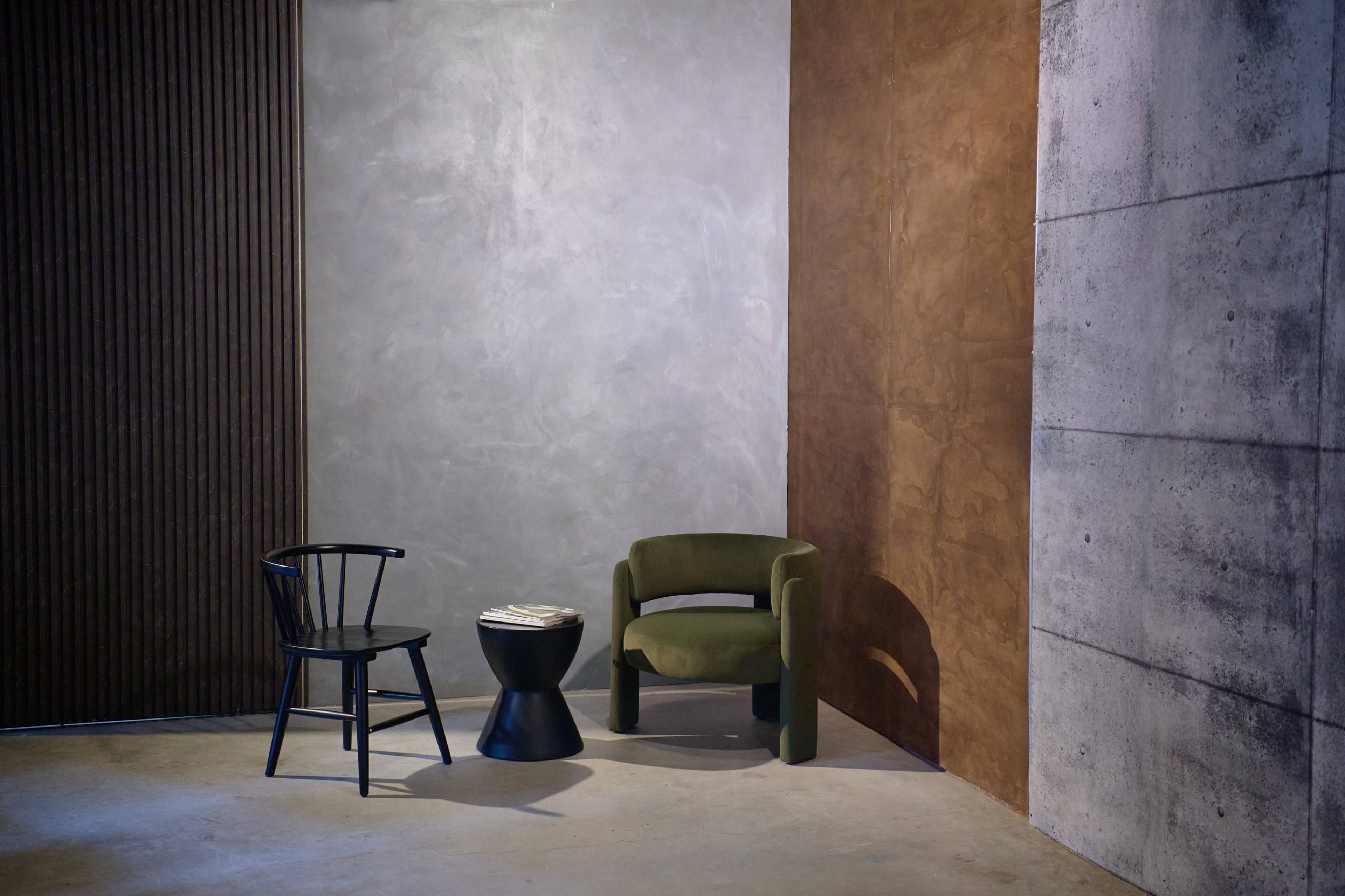 The image shows a minimalistic interior scene featuring a black wooden chair, a circular black side table, and a green upholstered armchair against textured walls in gray and brown tones.