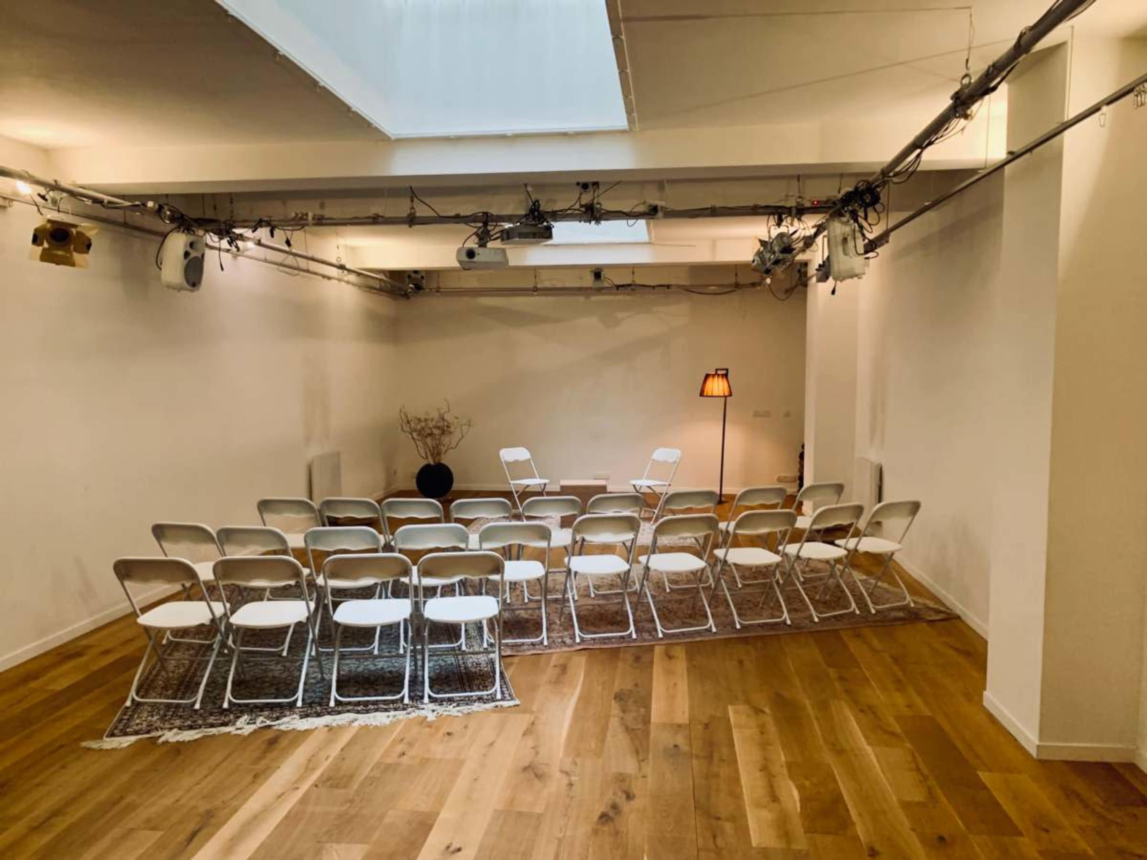The image shows a spacious room arranged with rows of white folding chairs facing a small decorative display and a lamp, on wooden flooring.