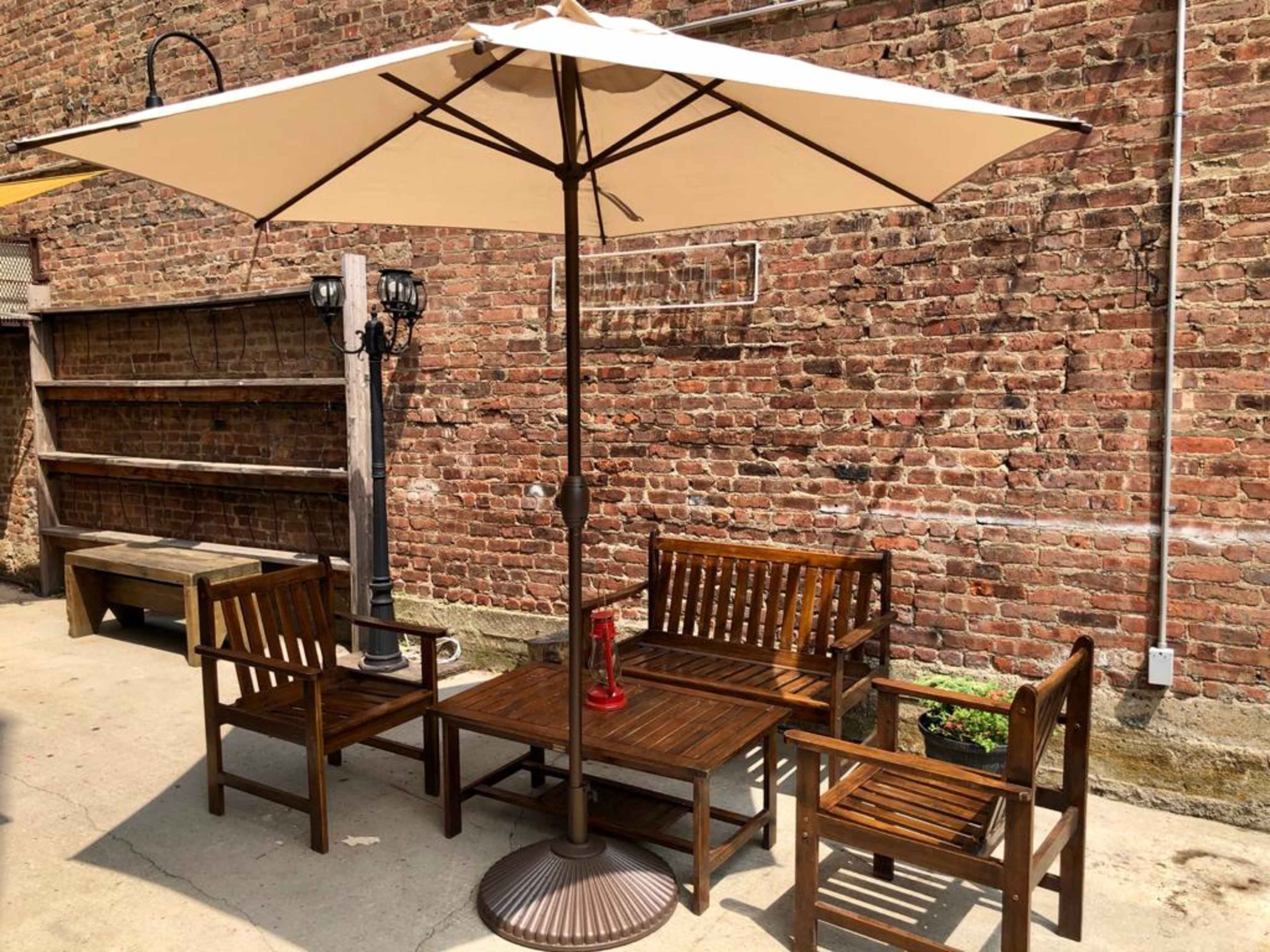 A wooden patio set with an umbrella is set against a brick wall, alongside a small lamp post and a shelf.
