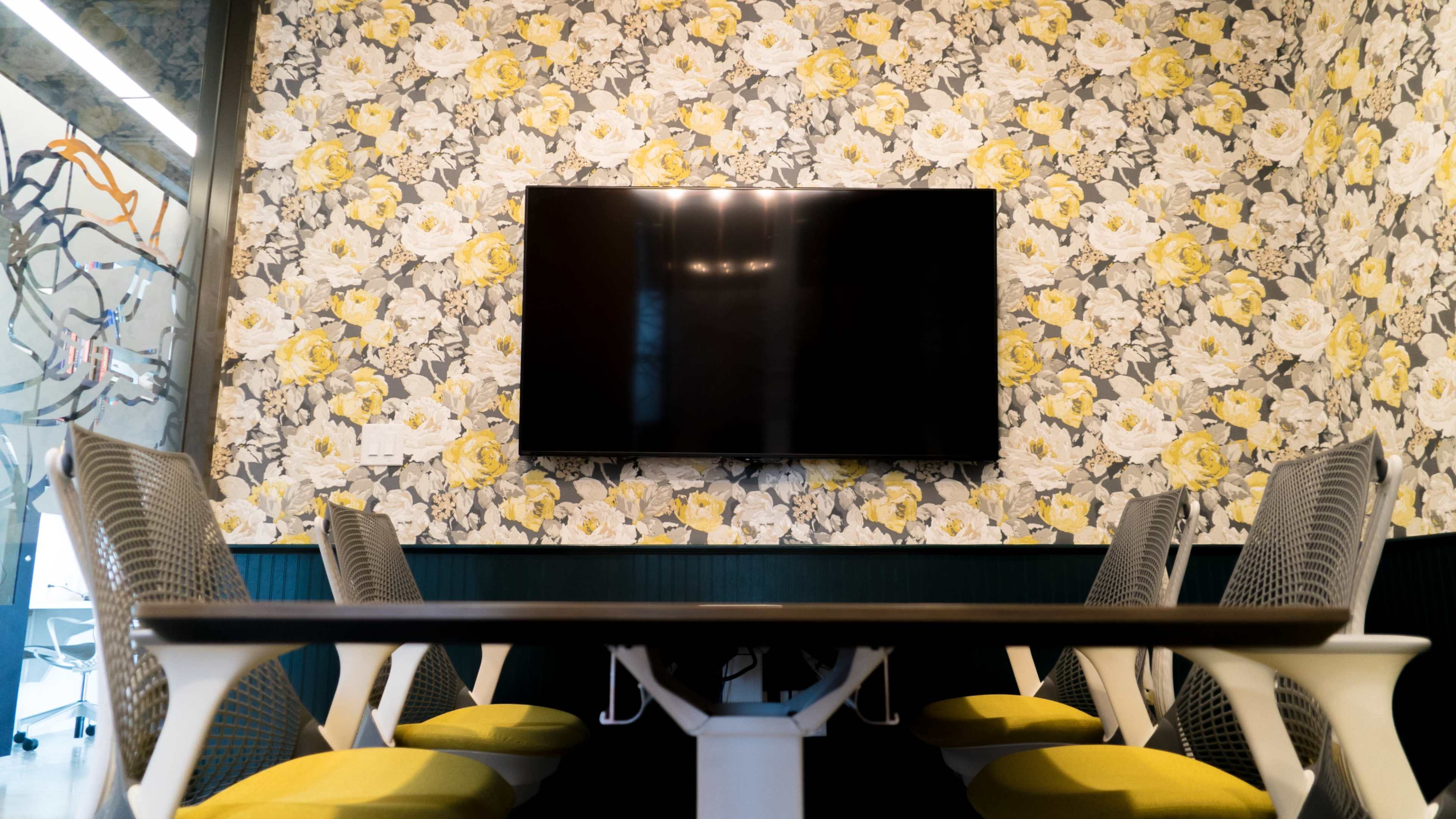 A conference room features a dark television mounted on a floral wallpaper and a long table surrounded by chairs.