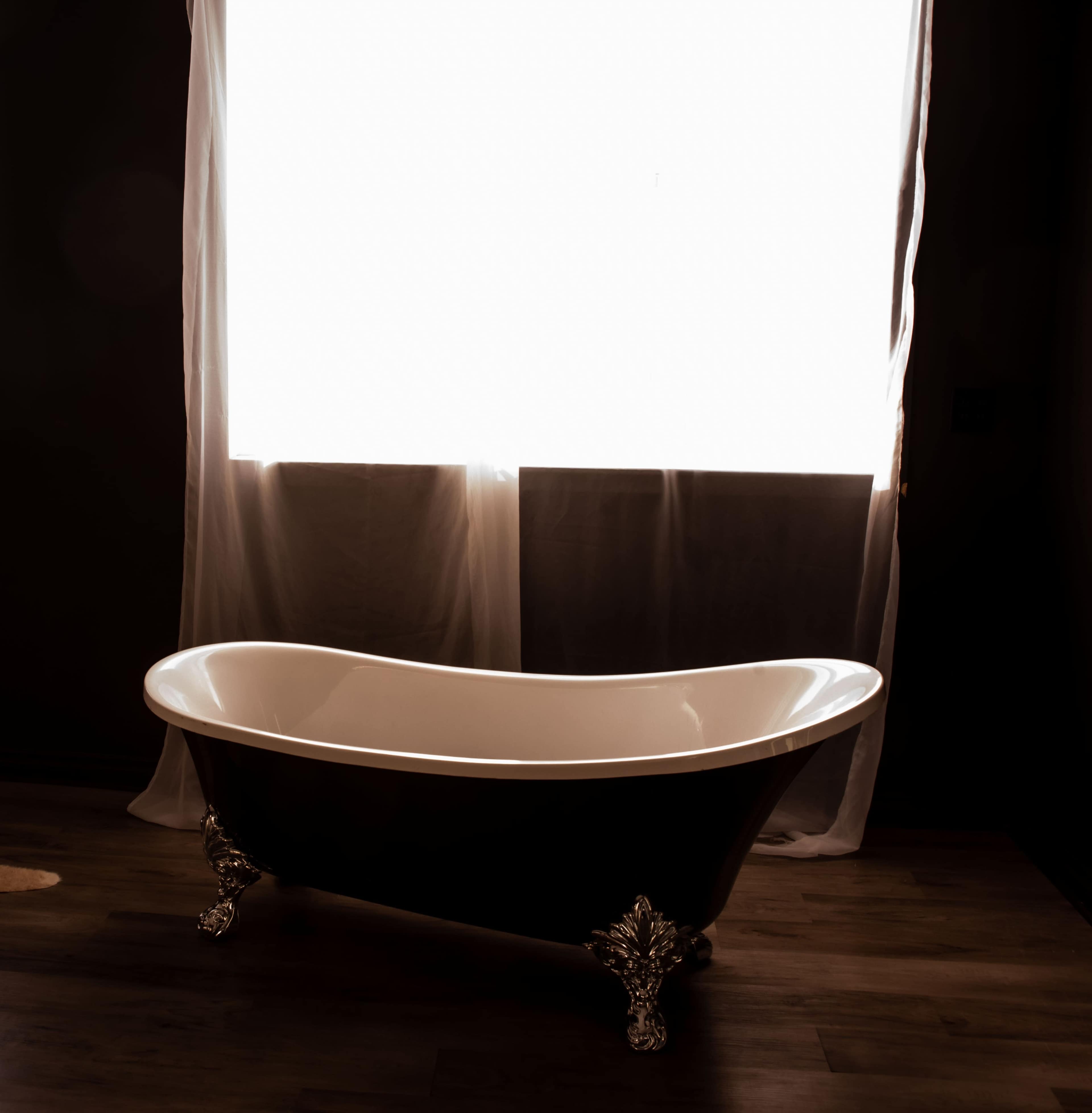 A freestanding black and white bathtub sits on wooden flooring beneath a bright, sheer curtain window.