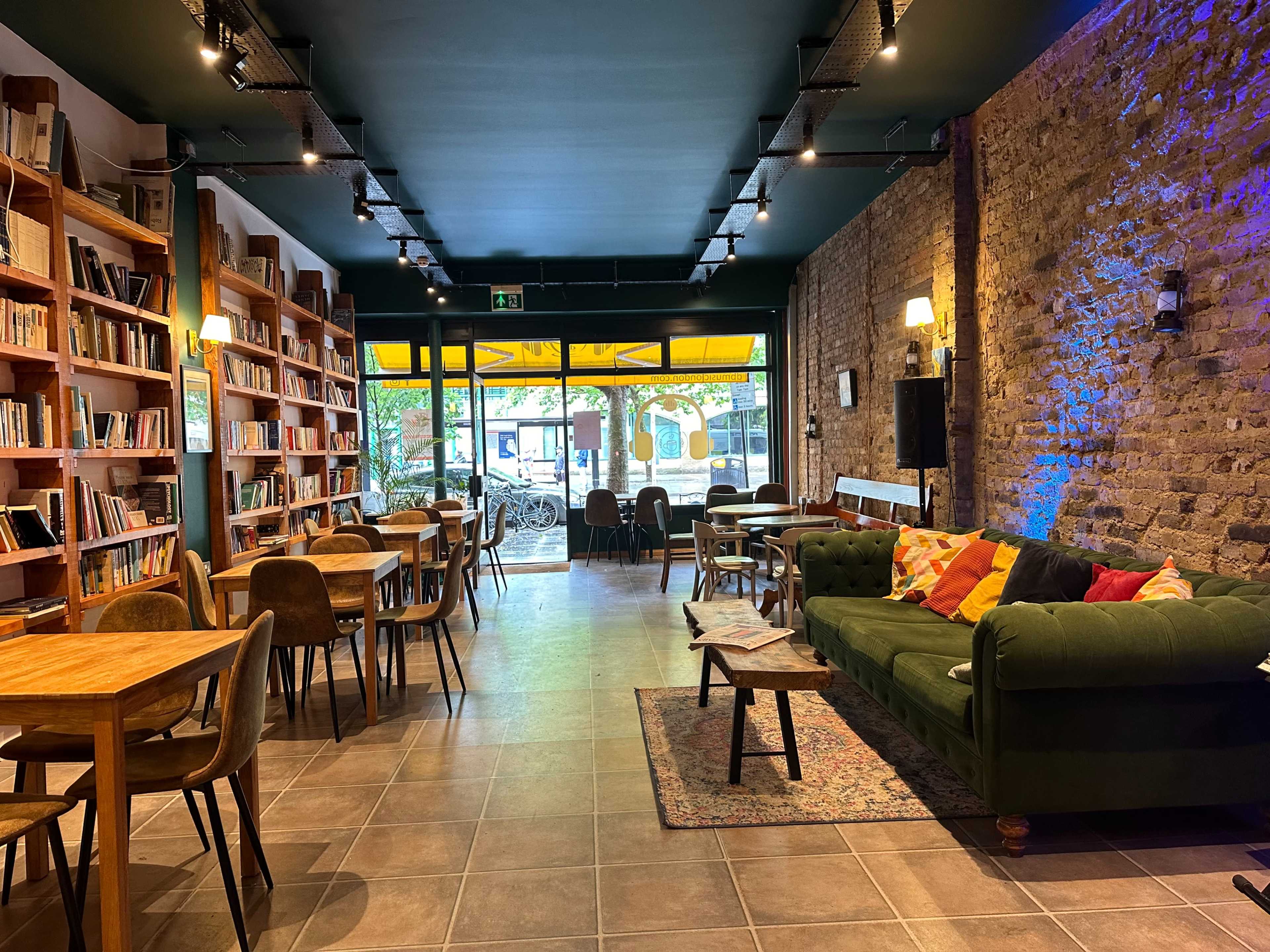 The image shows a cozy café interior with wooden tables, chairs, and a green couch, alongside shelves of books, and large windows overlooking a street.