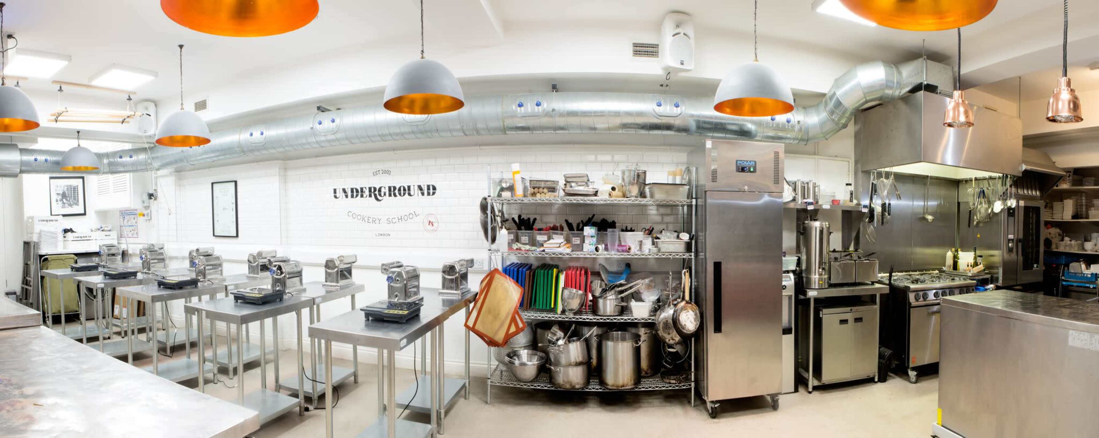 The image shows a modern kitchen with stainless steel appliances, cooking equipment, and organized workstations under bright overhead lights.