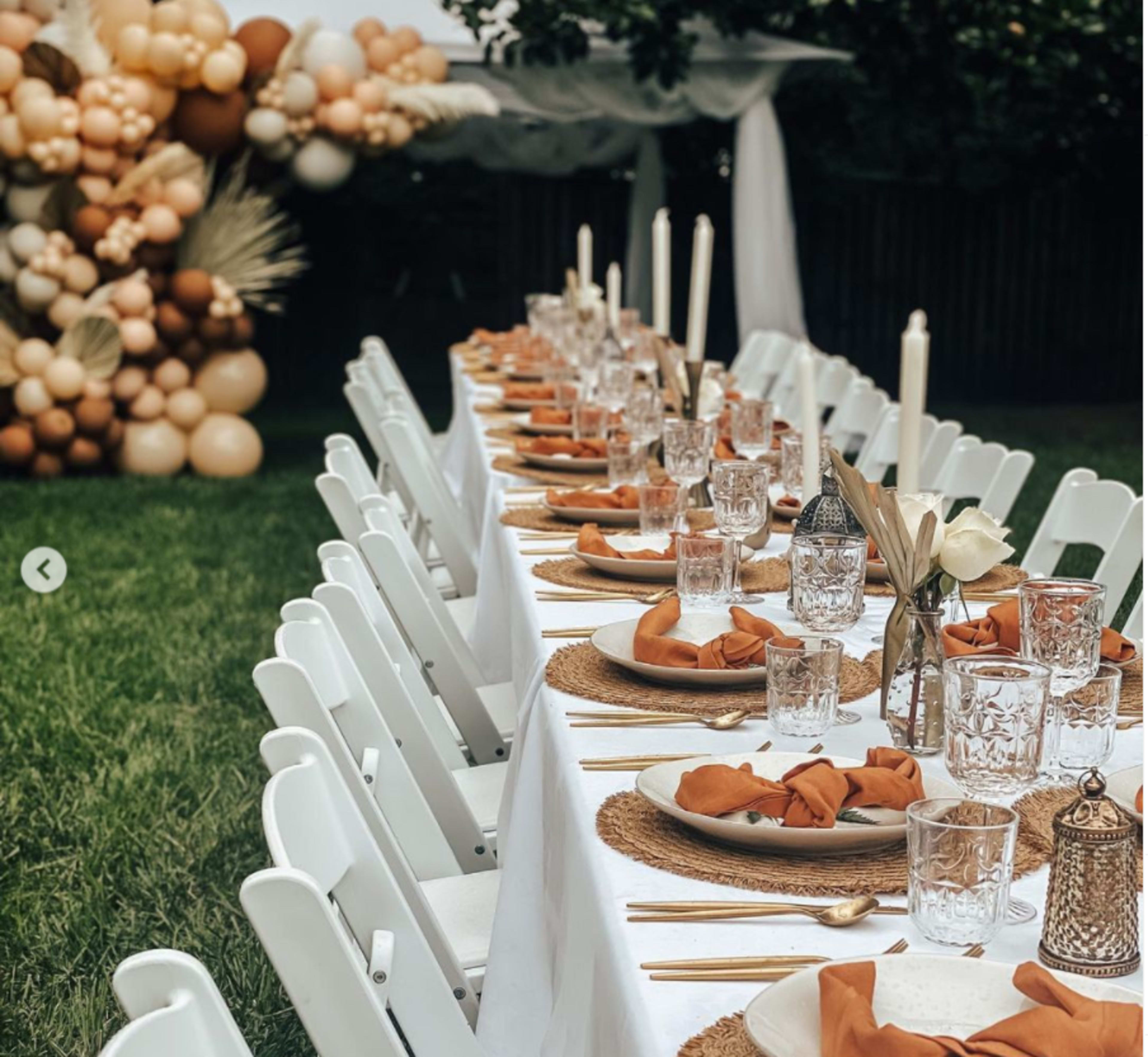 A long dining table is set outdoors with white chairs, decorated with plates, glasses, and orange napkins, in front of a balloon installation.