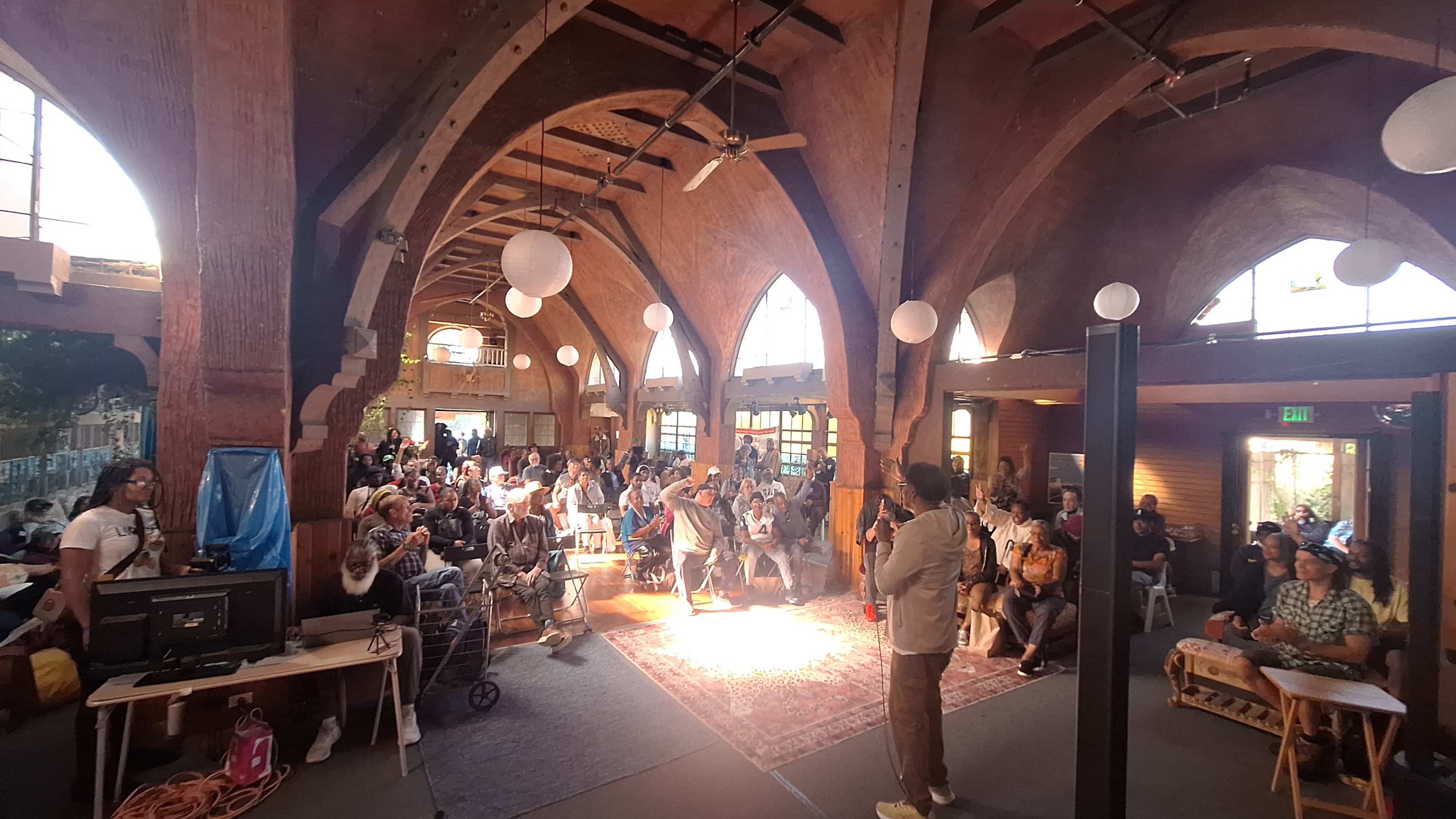A diverse audience sits in a spacious, arched room with wood beams, while a speaker stands on a rug at the center, engaging the crowd.