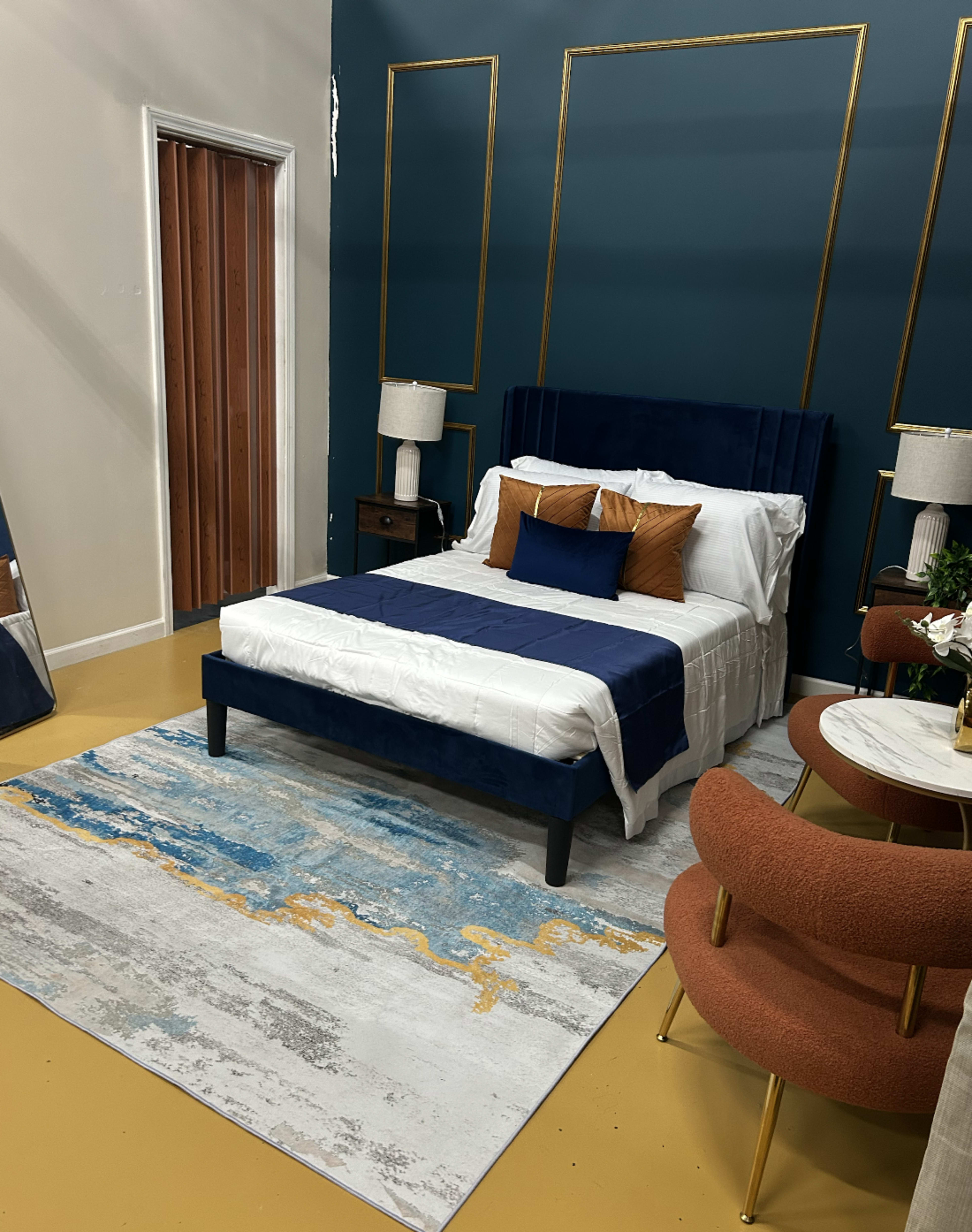 The image shows a beautifully staged bedroom featuring a bed with a navy blue headboard, decorative pillows, and a blue and gold area rug, accompanied by two orange chairs and a small side table.