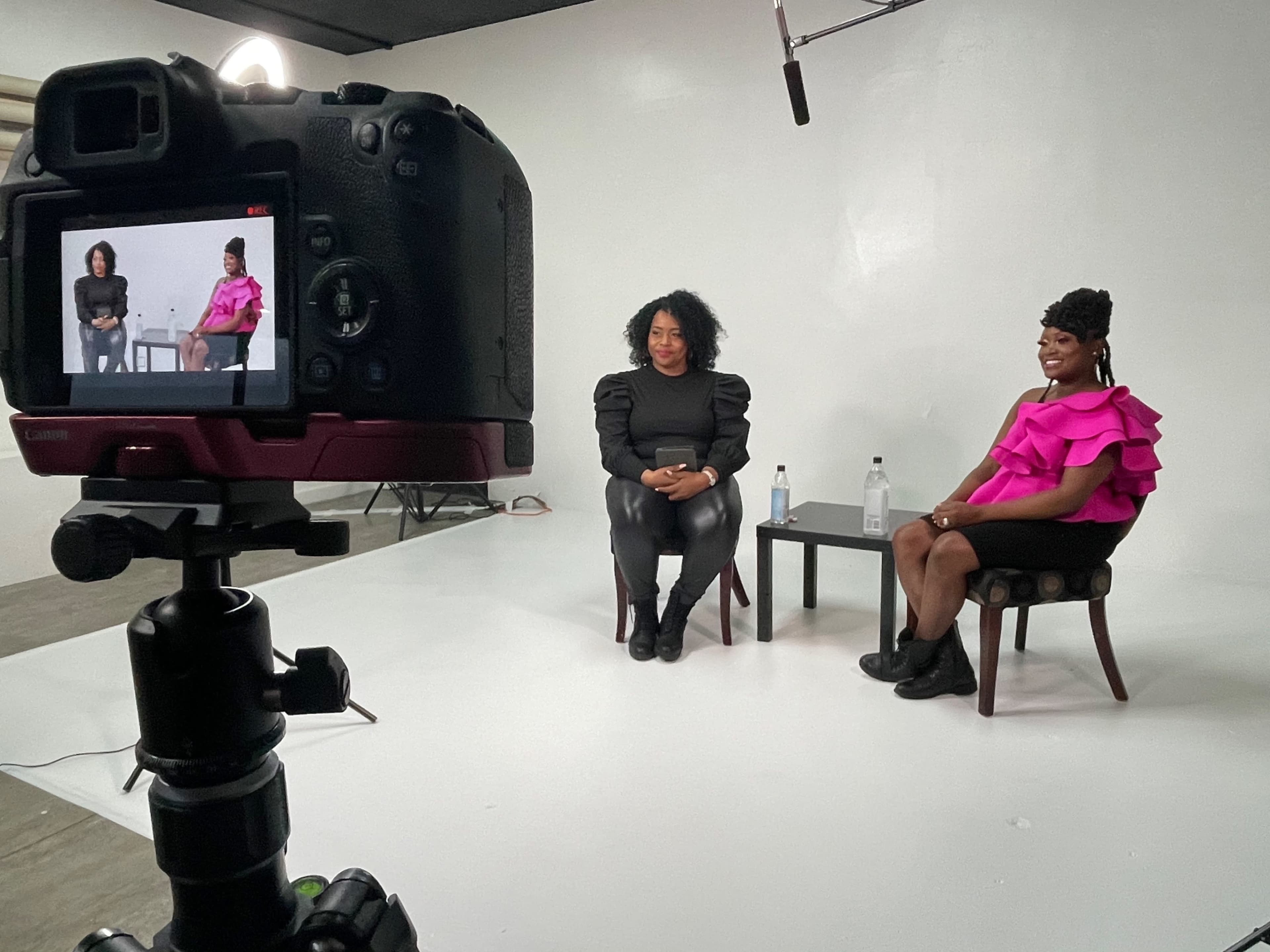 Two women sit on chairs in a studio while being filmed by a camera on a tripod.