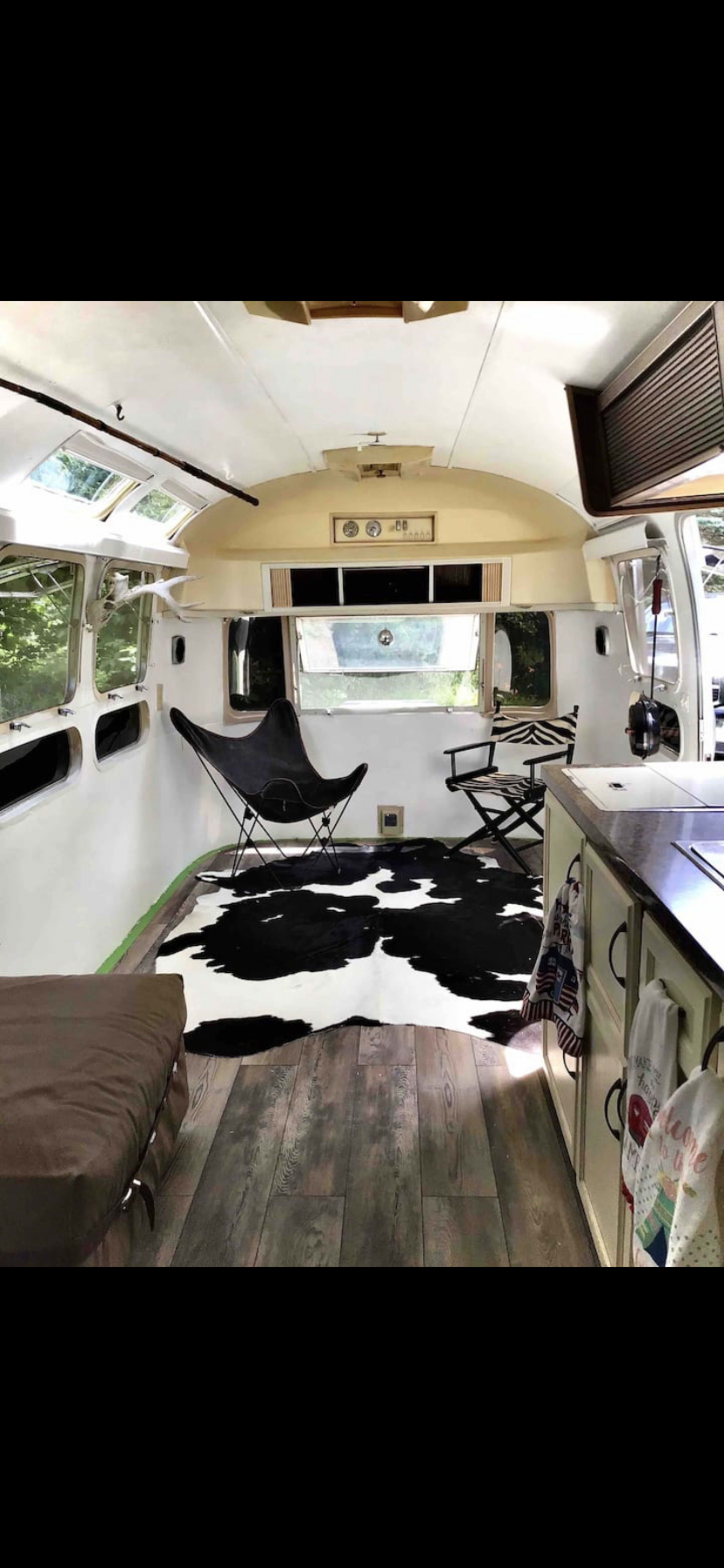 The interior of a vintage Airstream featuring a black and white cowhide rug, a folding chair, and a sectional couch.