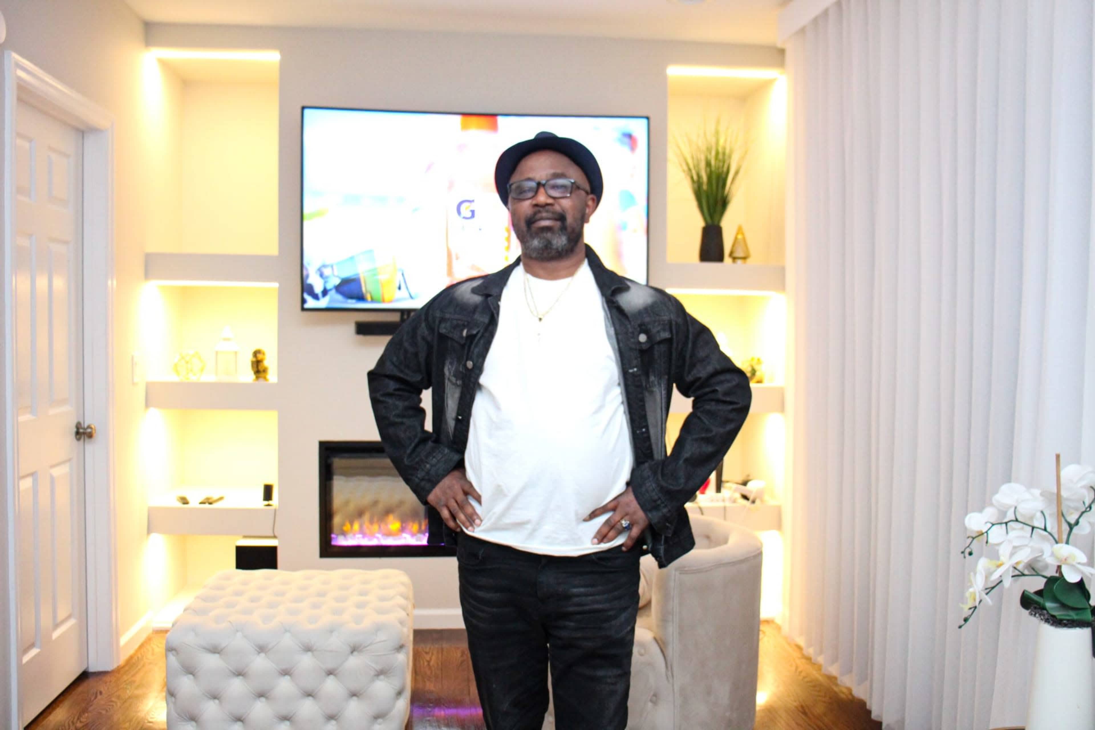 A man stands with his hands on his hips in a modern living room featuring a television and a decorative fireplace.