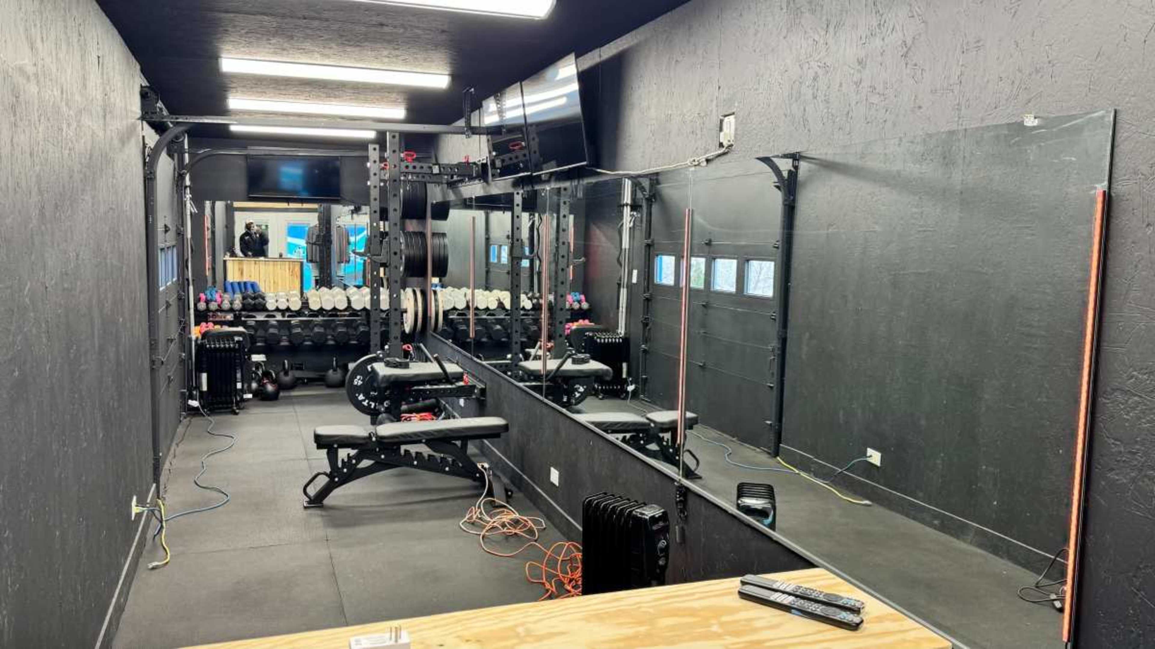 The image shows a gym space equipped with weightlifting benches, dumbbells, and mirrors along the walls.