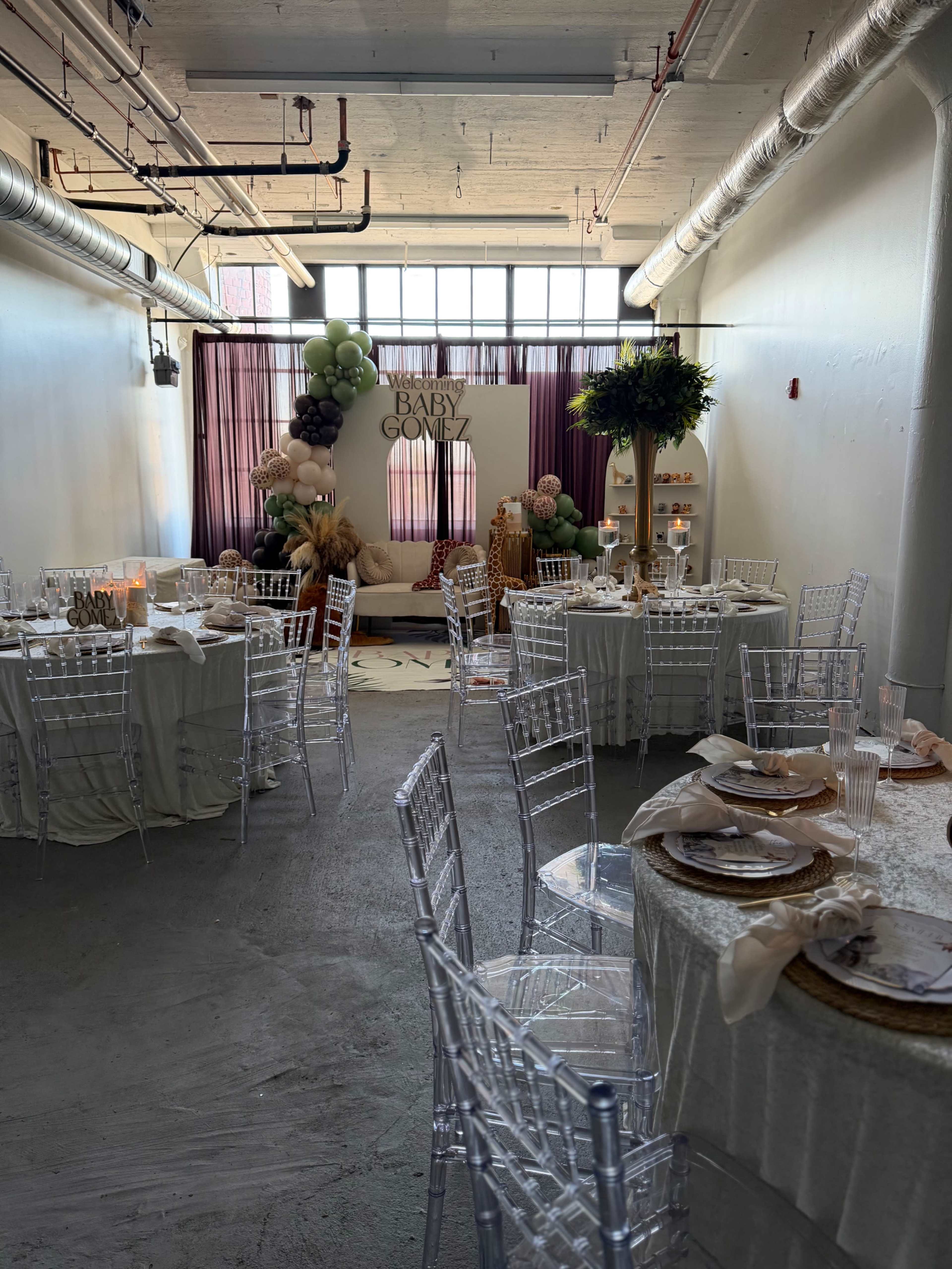 The image shows a decorated event space featuring several tables with clear chairs, a backdrop with the words "Baby Gomez," and various decorative elements, including balloons and plants.