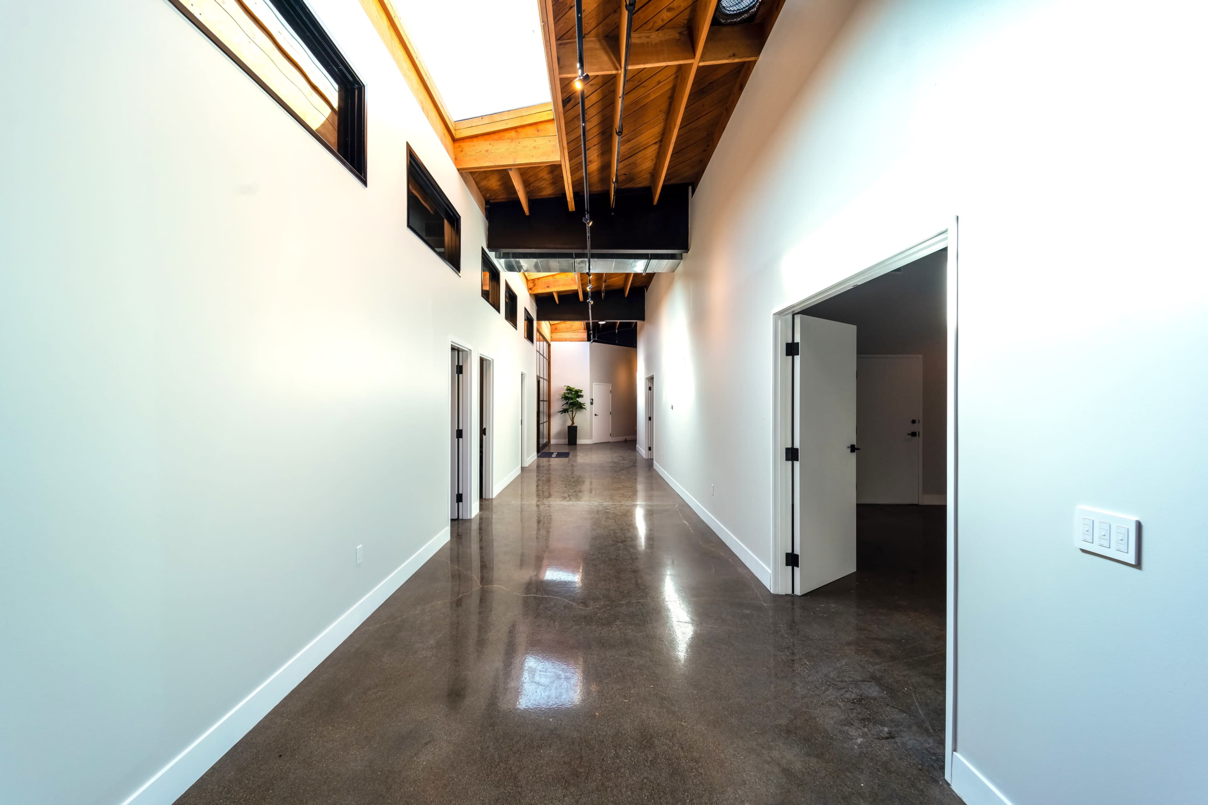 The image shows a modern hallway with polished concrete floors, white walls, and wooden beams overhead, lined with several doors on either side.