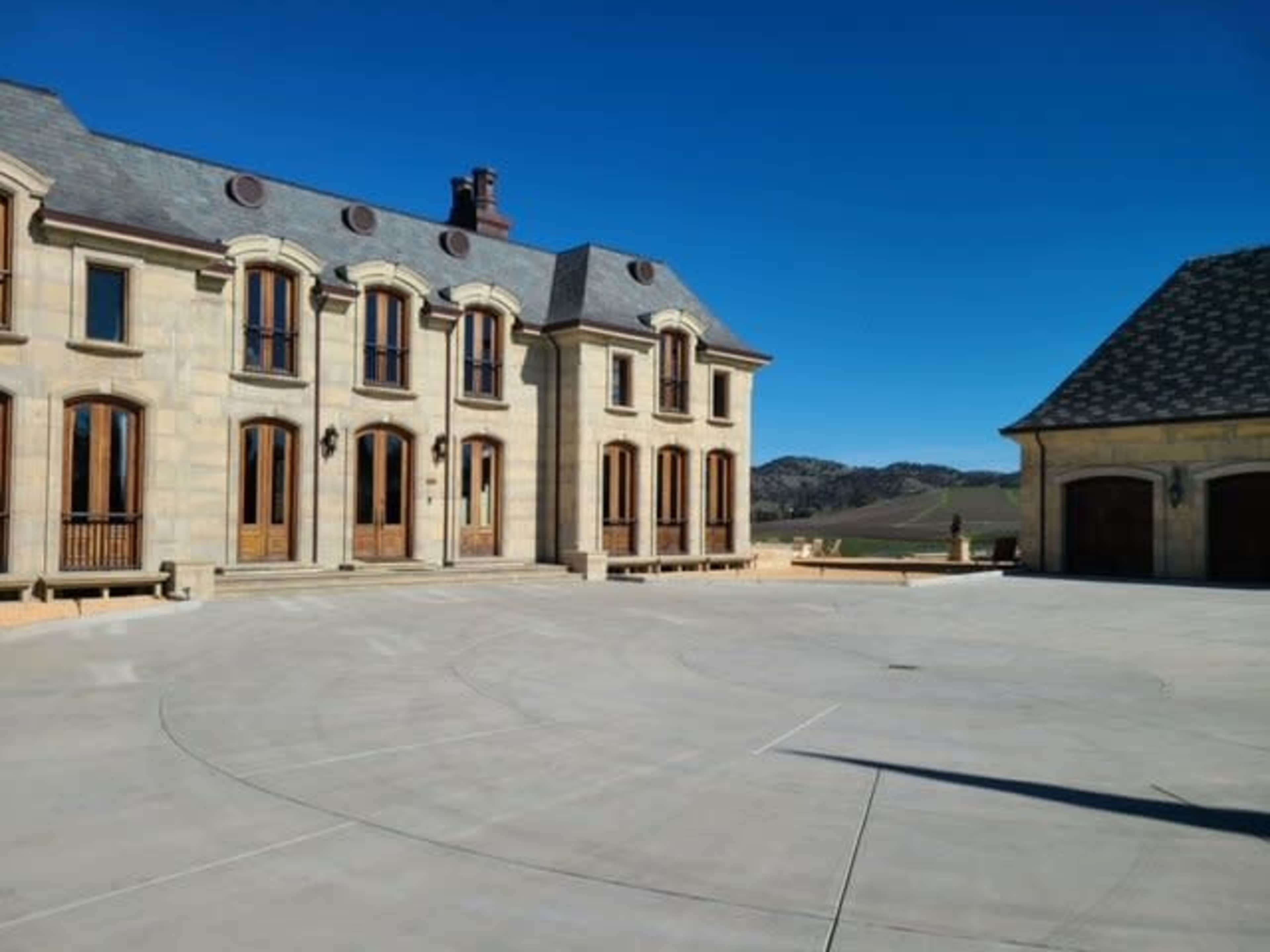 A large, stone mansion features multiple arched windows and a wide concrete courtyard under a clear blue sky.