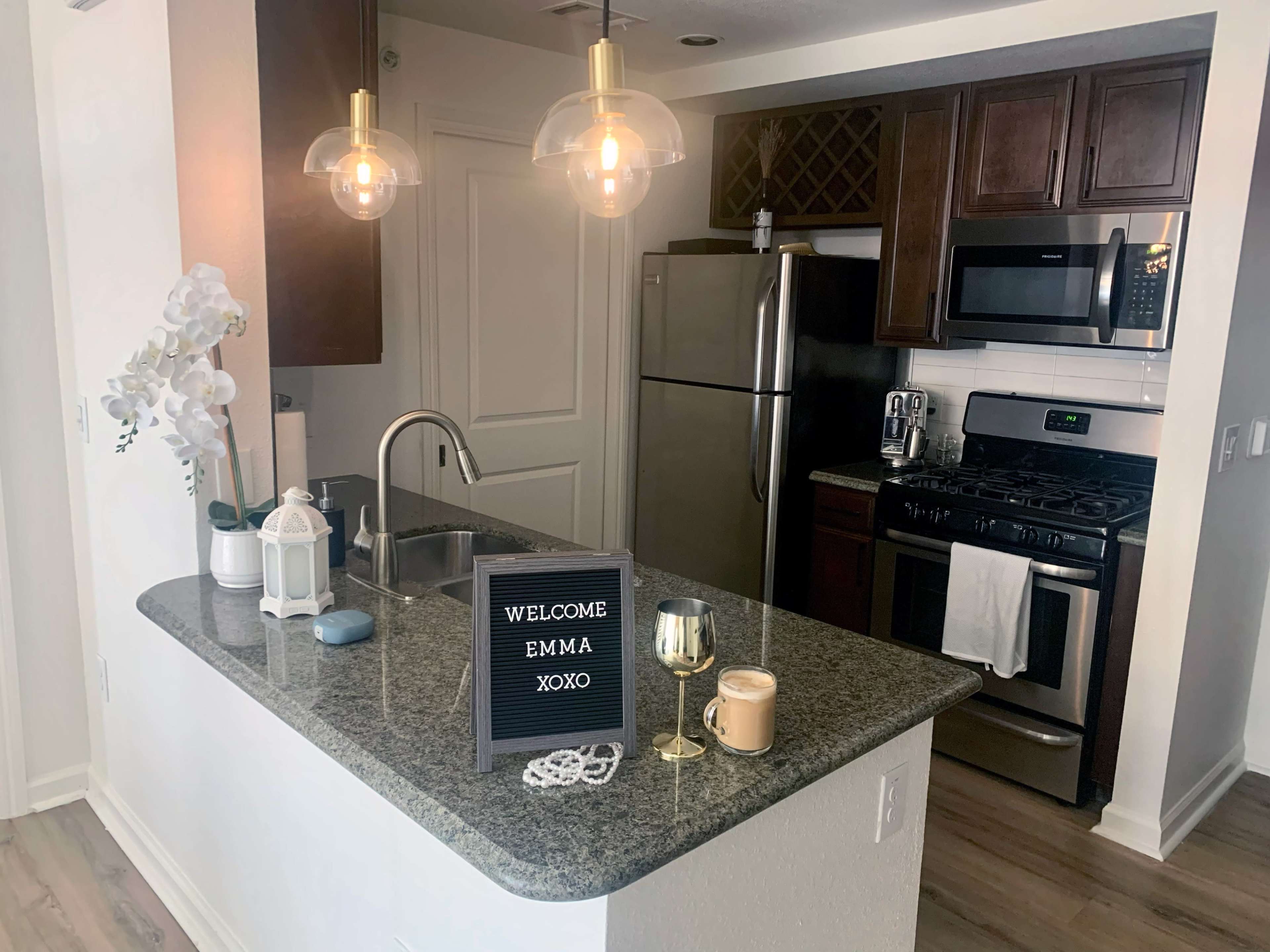 A modern kitchen features a granite countertop, stainless steel appliances, pendant lights, and a welcome sign.
