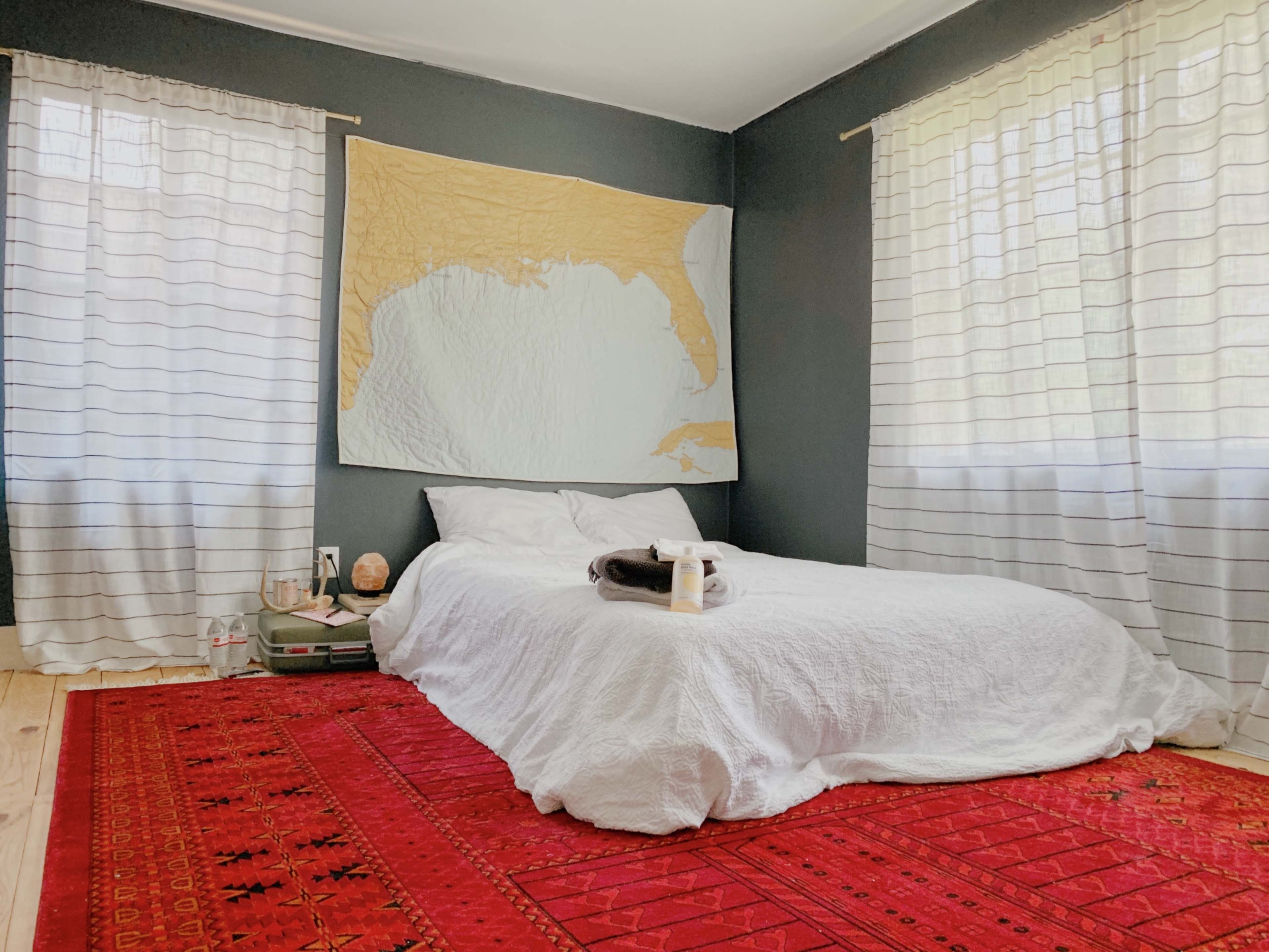 A simple bedroom features a white bedspread on a bed, with a large map hanging on the wall, flanked by light-colored curtains and a red area rug.