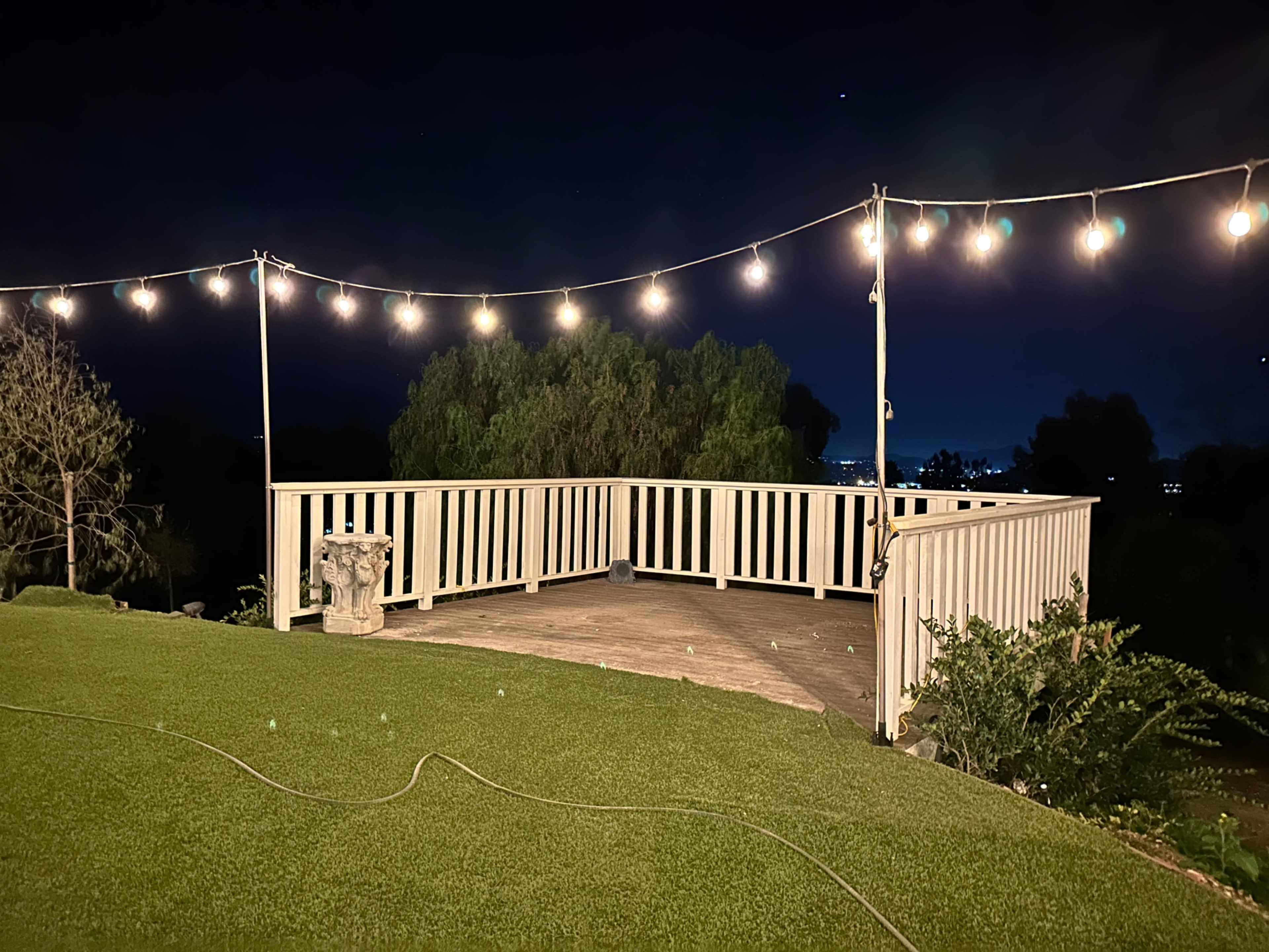 A wooden deck with string lights overhead and a grassy area in the foreground, set against a night sky.