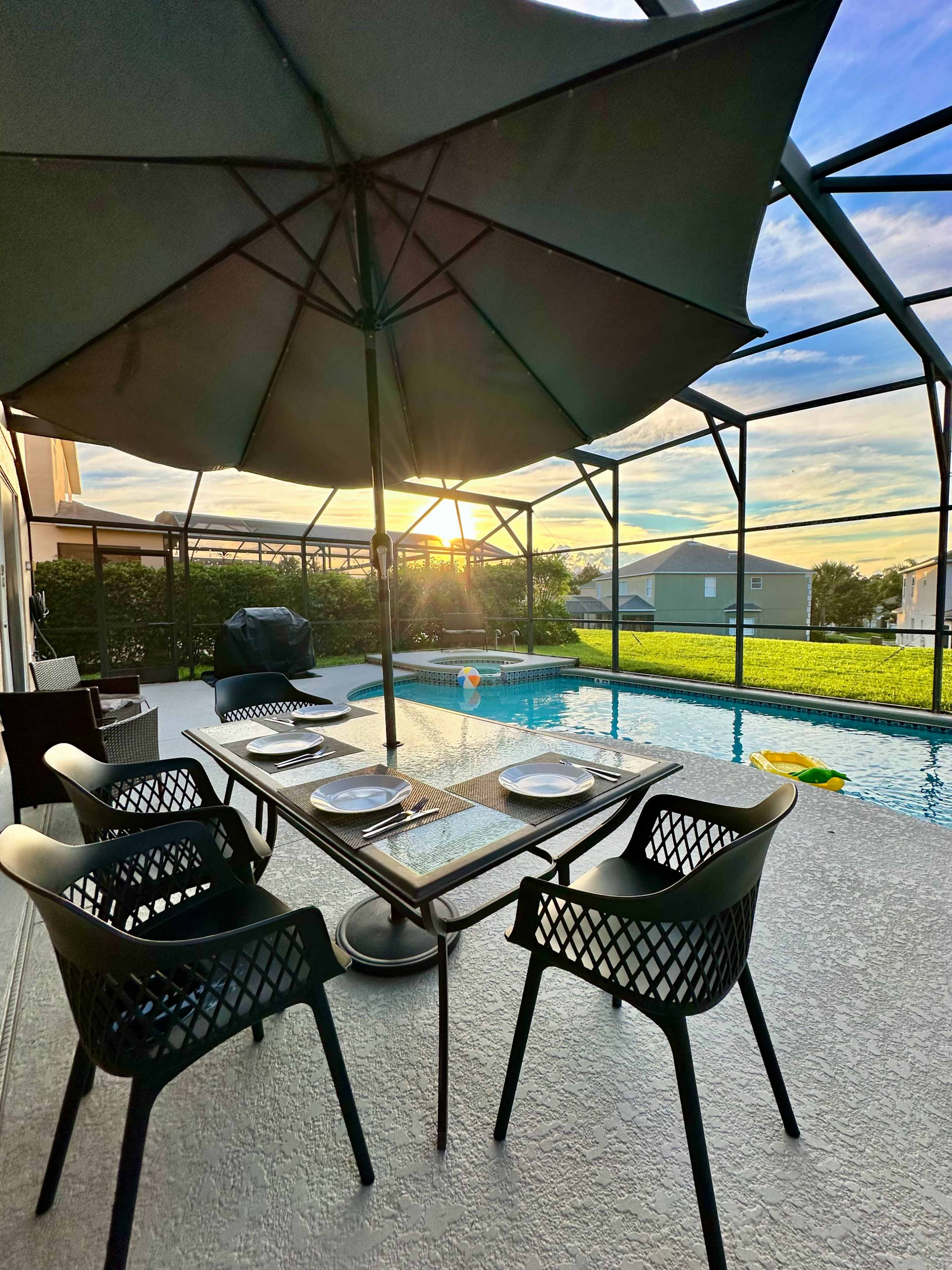 A patio dining area with black chairs and a table is set beside a swimming pool, with a sun setting in the background.