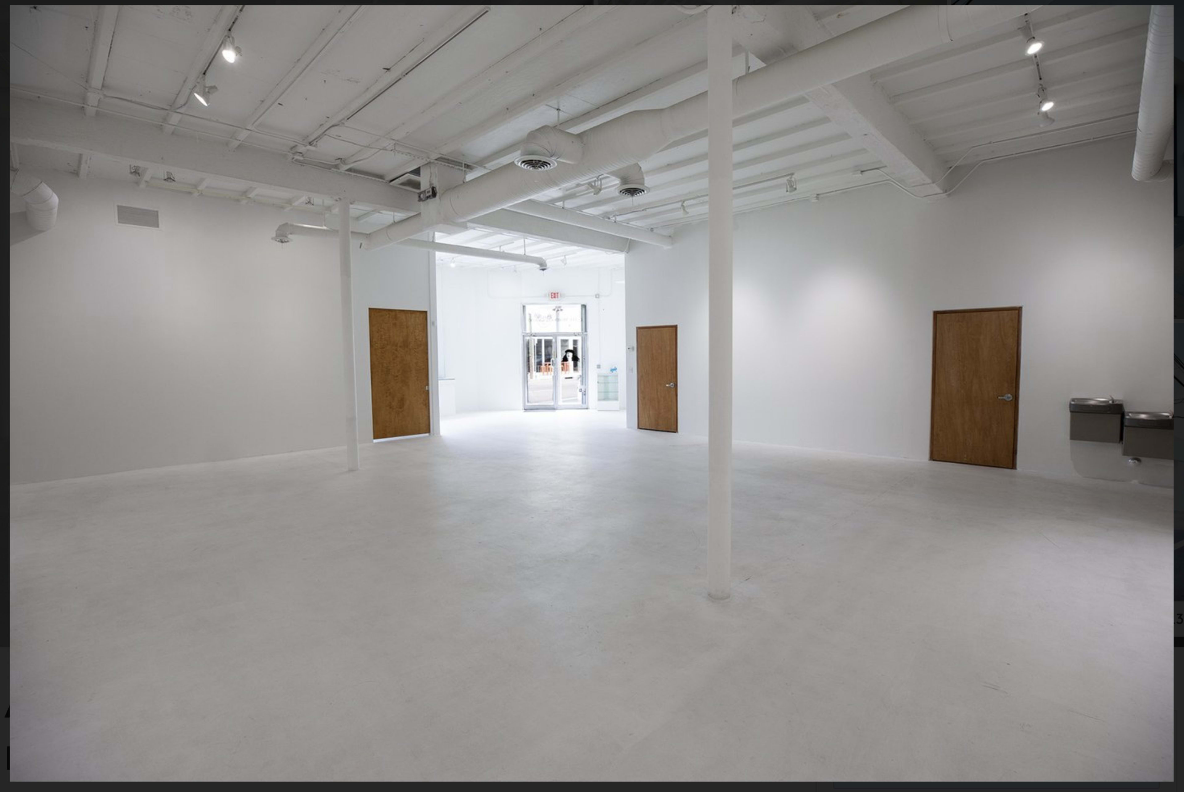 The image shows a spacious, empty room with white walls and a concrete floor, featuring two wooden doors and an entrance leading outside.