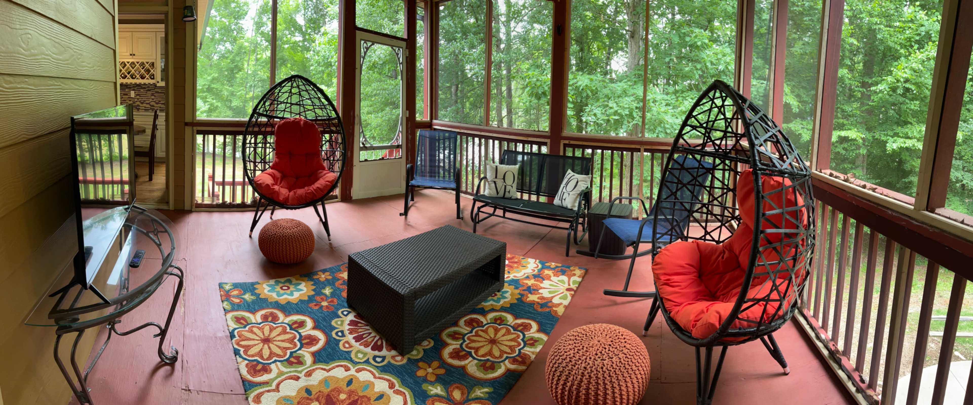 A screened-in porch featuring two hanging chairs with red cushions, a black coffee table, a fabric couch, and a colorful patterned rug, surrounded by greenery outside.