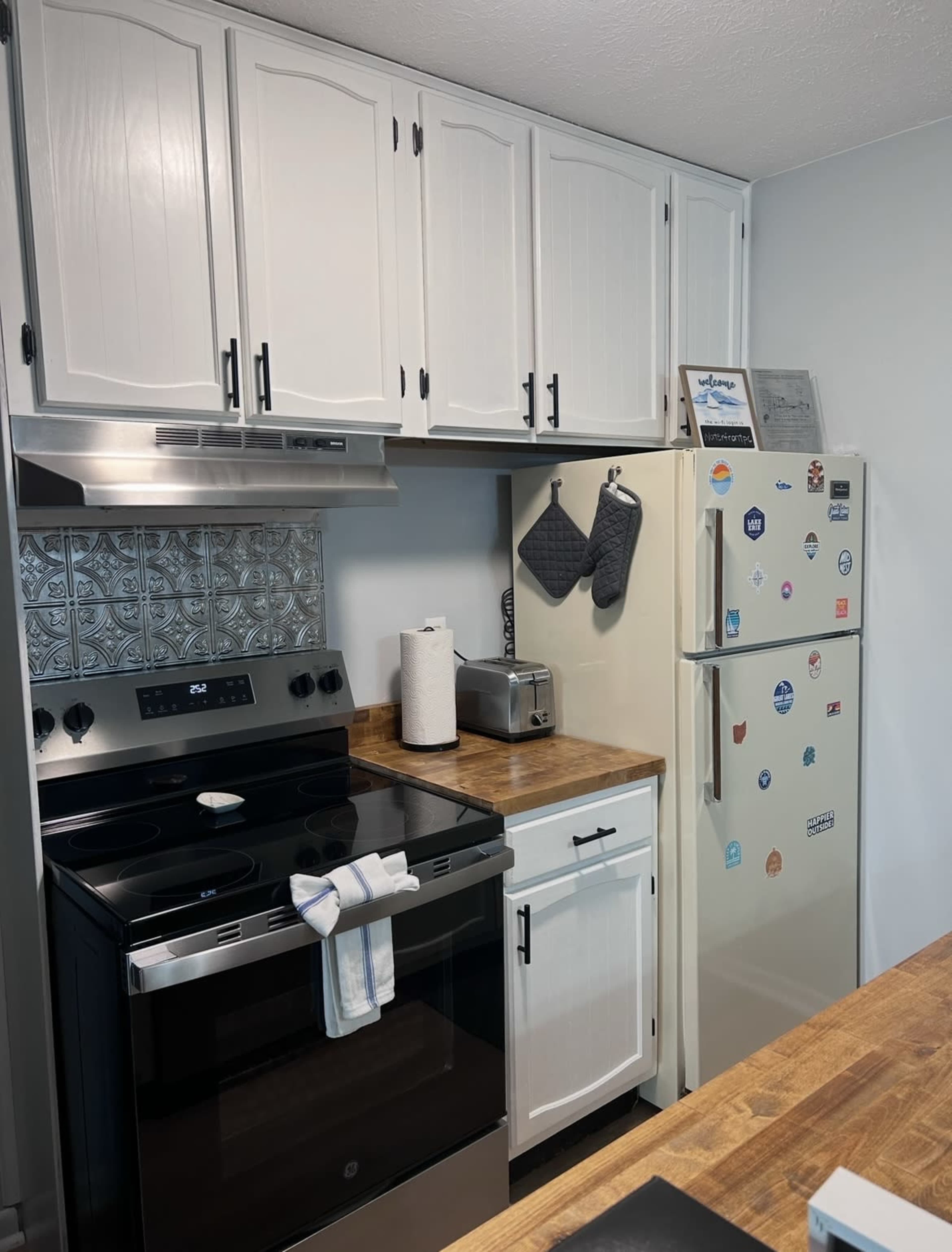 The image shows a modern kitchen with white cabinets, a black stove, a stainless steel refrigerator, and a wooden countertop.