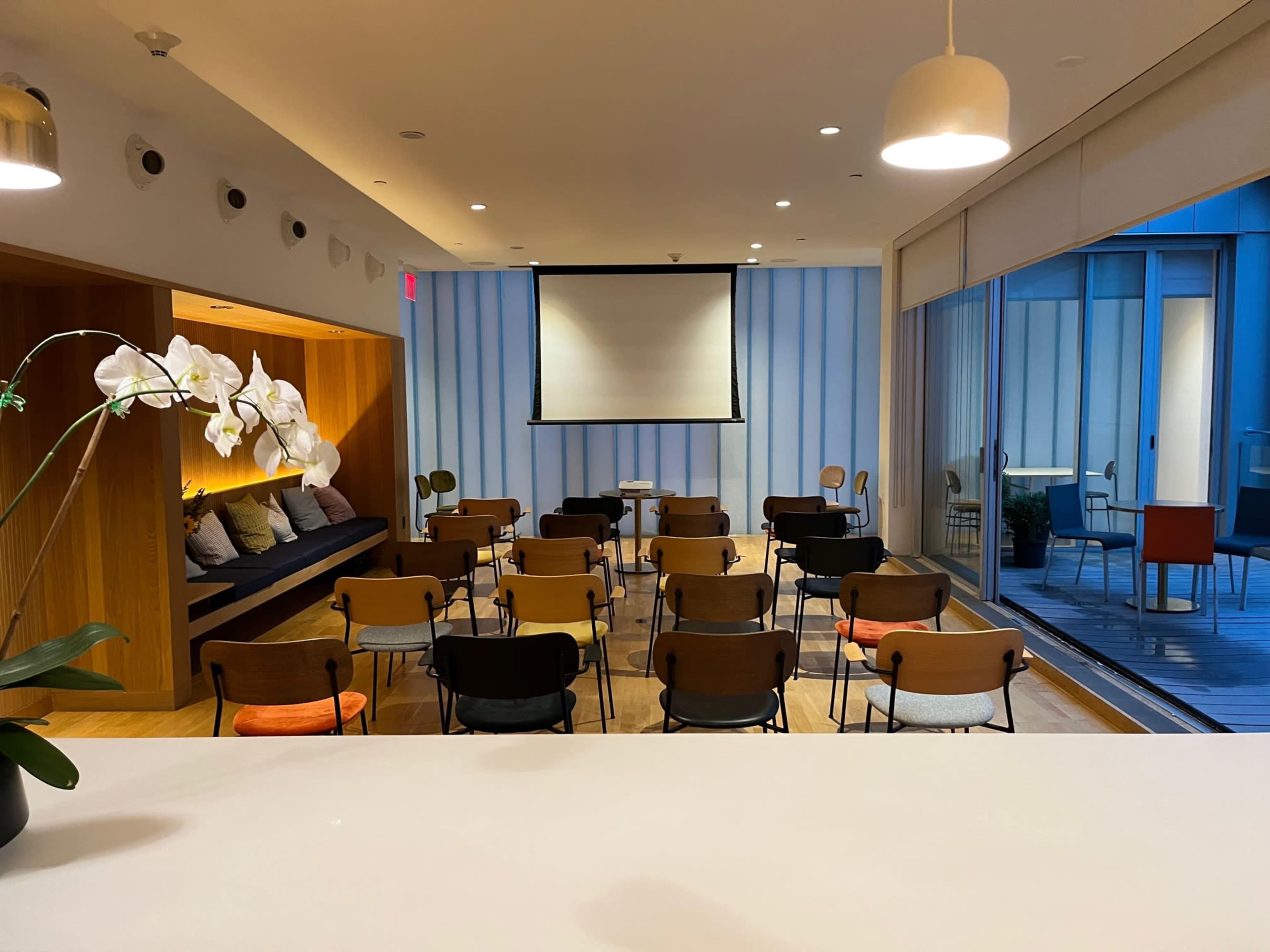 A modern seminar room set up with rows of chairs facing a projection screen, with a seating area and large windows on one side.