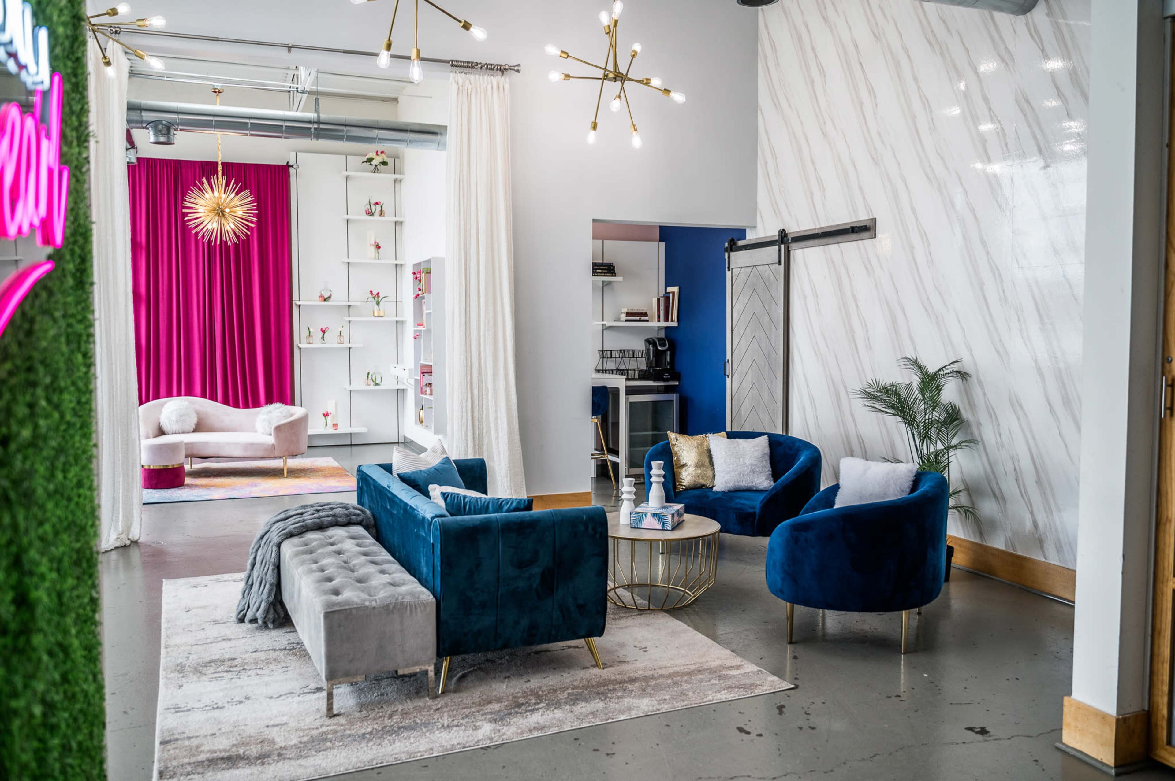 A modern interior space featuring a seating area with blue velvet chairs and a gray tufted sofa, accentuated by decorative lighting and a vibrant backdrop.
