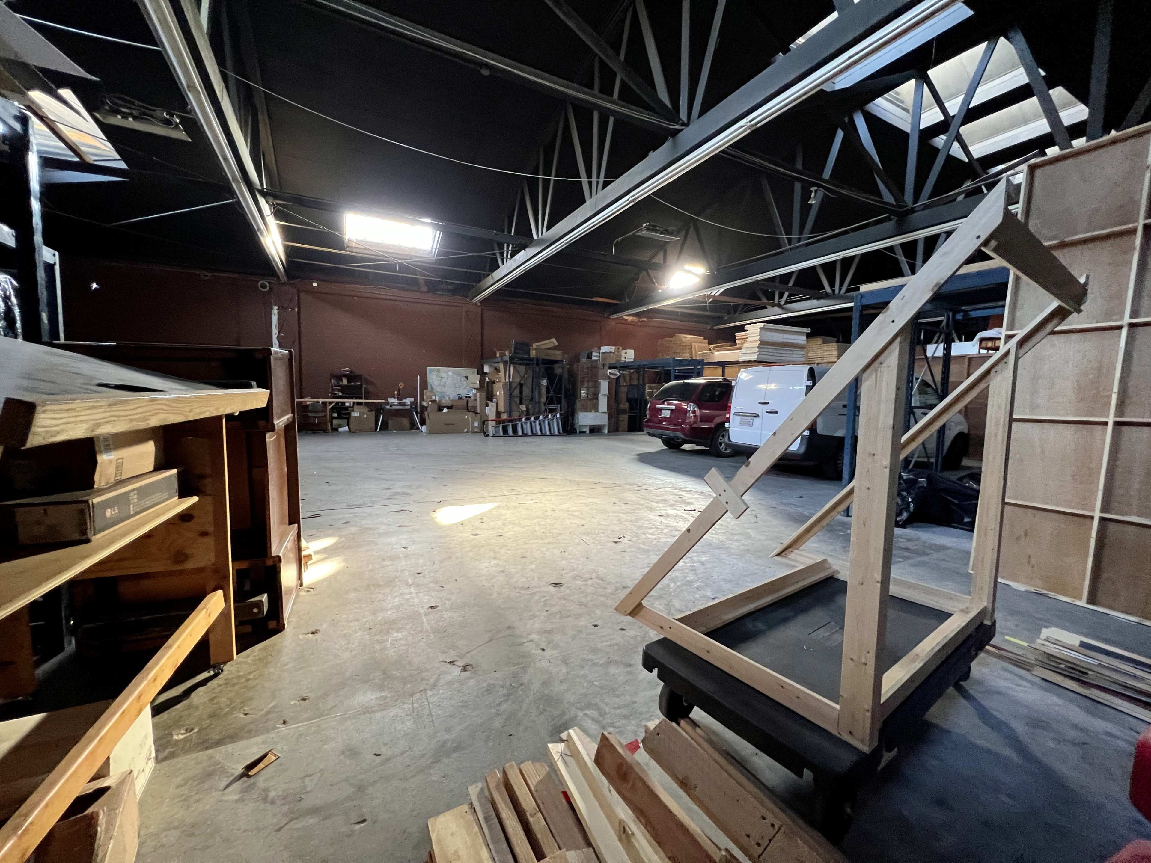 An empty warehouse interior features wooden storage pallets, a cart with a frame, and various stacked boxes in the background.