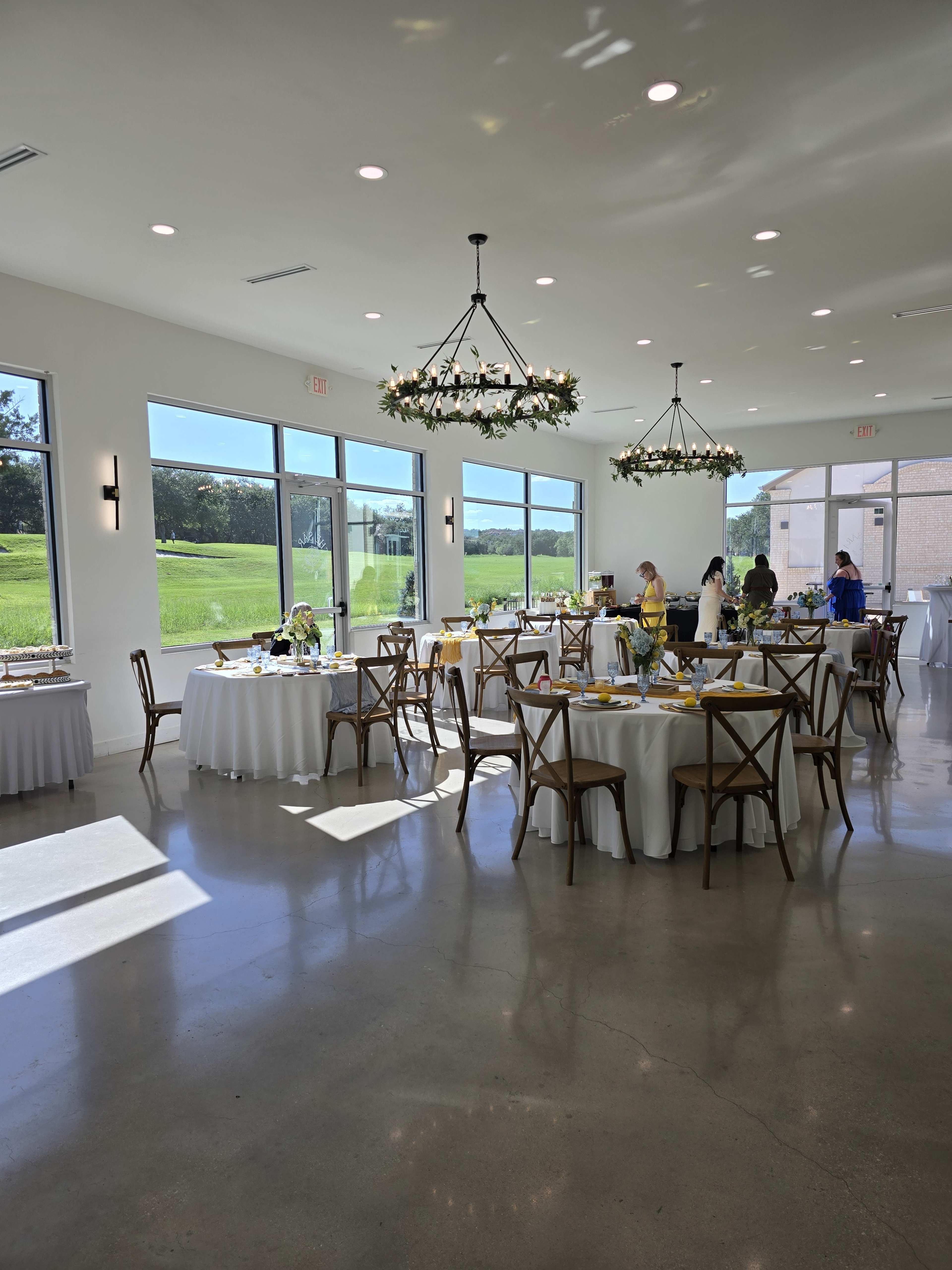 A bright event space features several round tables set with white linens, surrounded by wooden chairs, as people arrange decorations and prepare for a gathering.