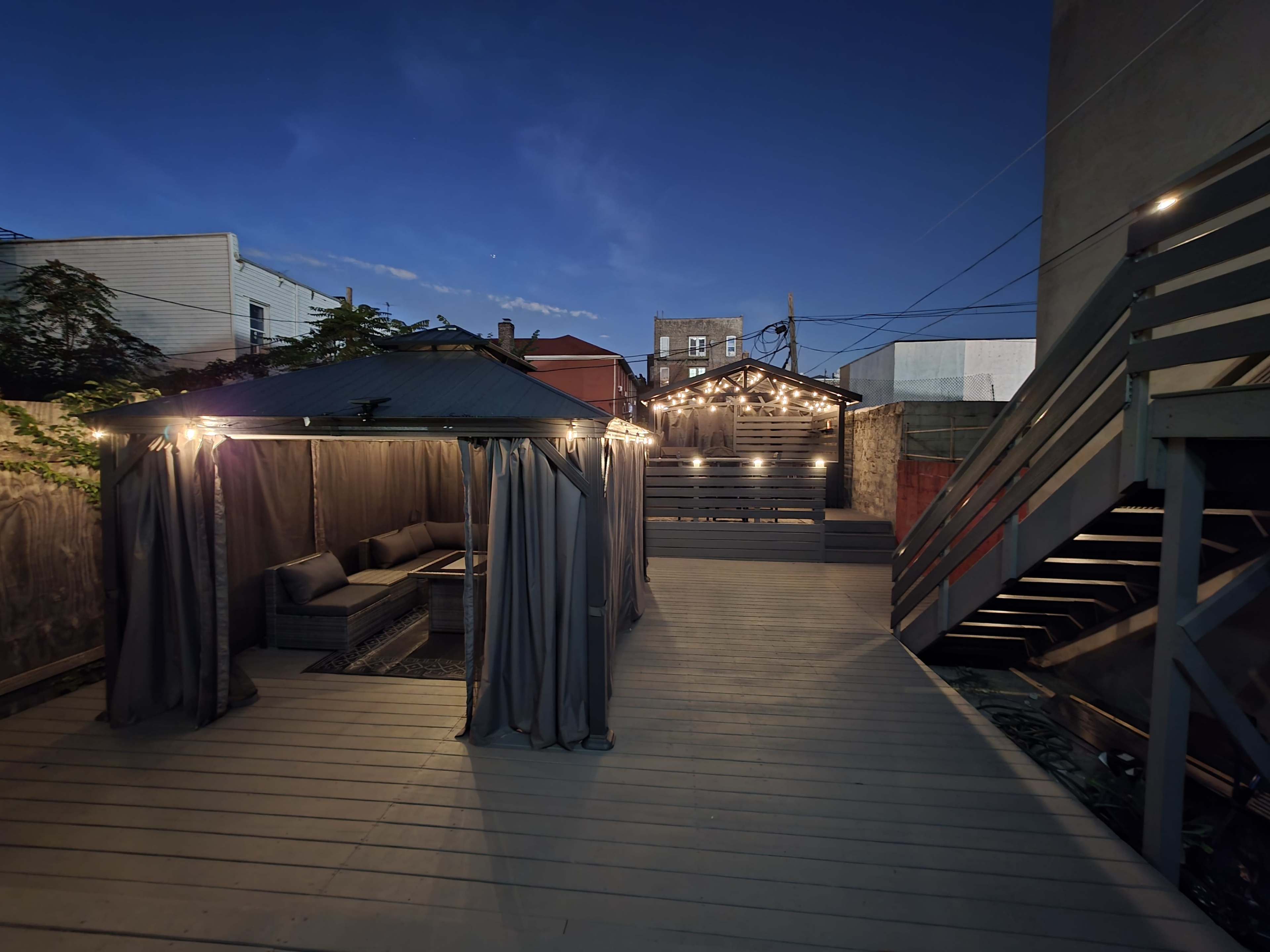 The image shows a wooden deck area with a covered seating space, string lights, and a neighboring building in the background under a twilight sky.