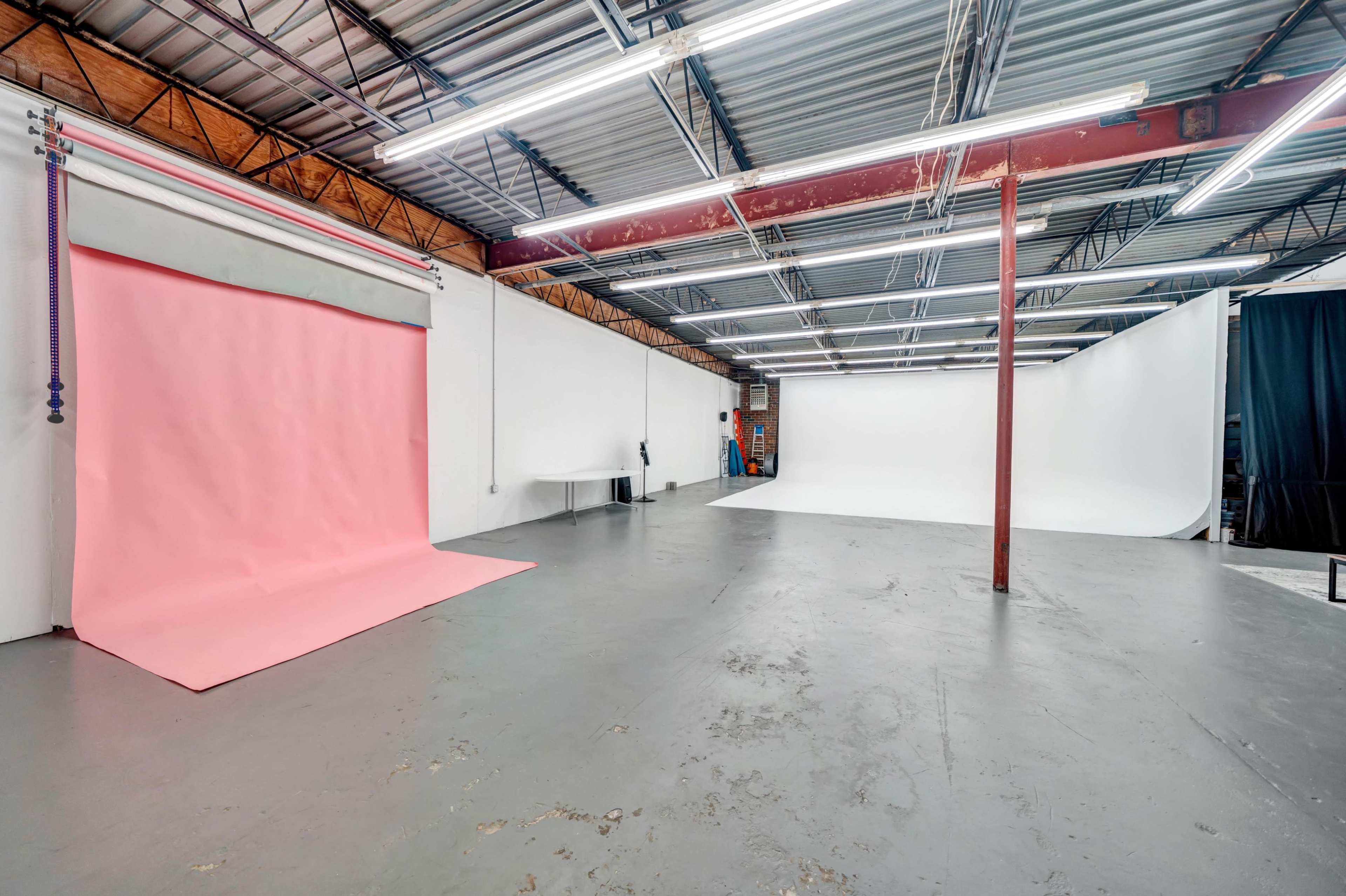 A spacious photography studio featuring two backdrop areas, one with a pink backdrop and another with a white backdrop, along with overhead fluorescent lighting.