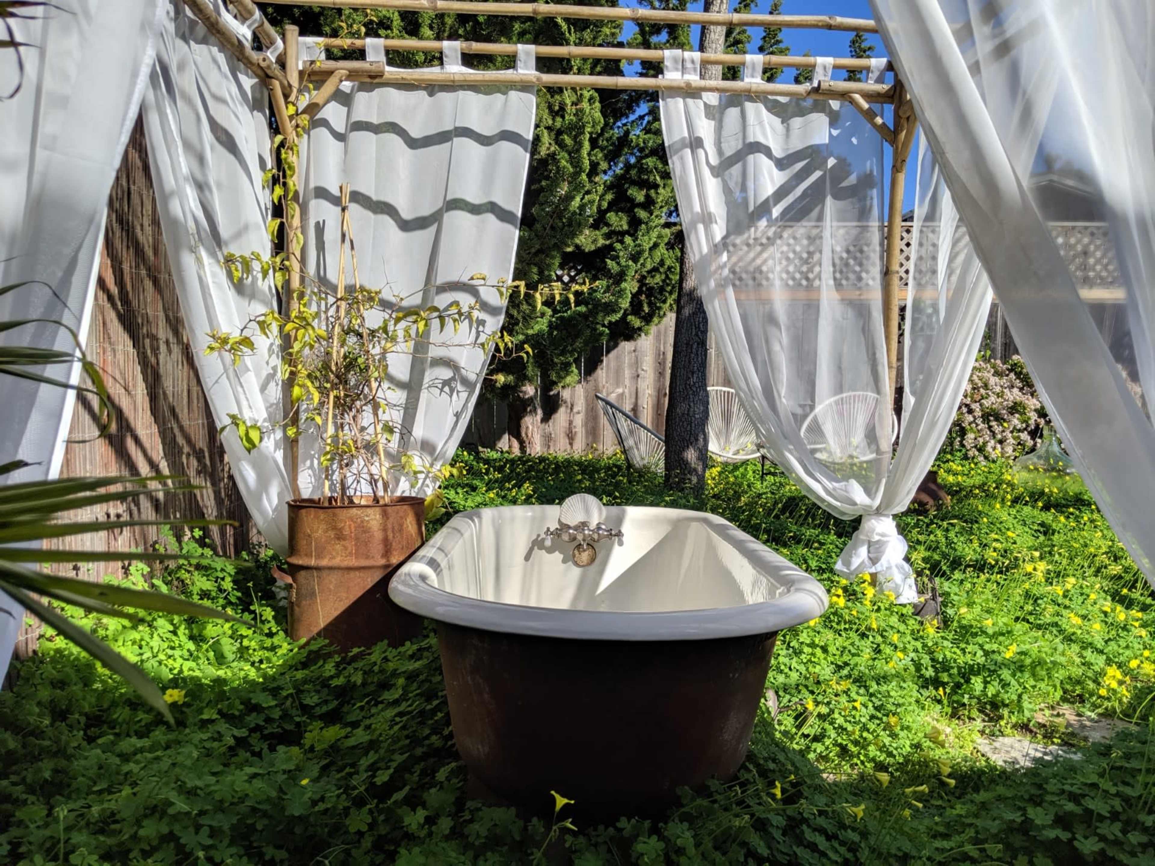 A vintage bathtub is positioned in a lush garden surrounded by sheer drapes and greenery.