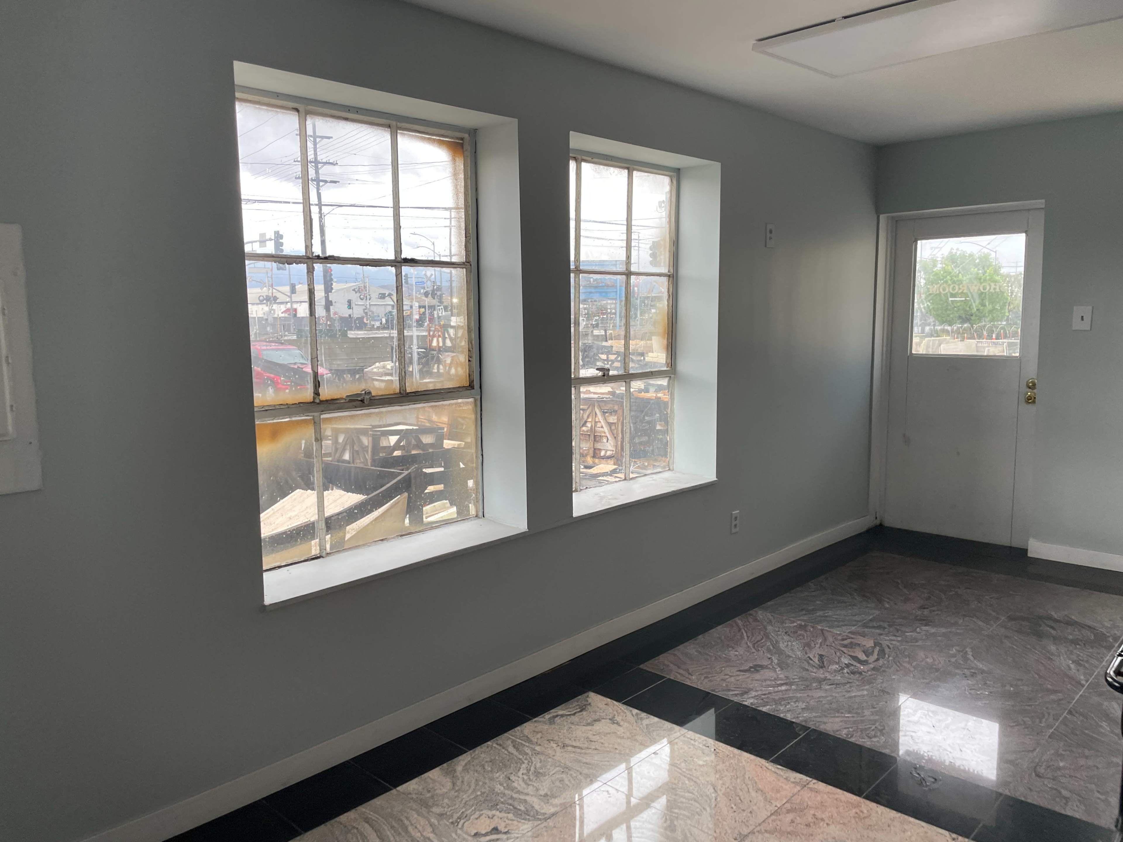 The image shows a room with two large windows on one wall, revealing an outdoor view, and a door on the right side leading to another area.