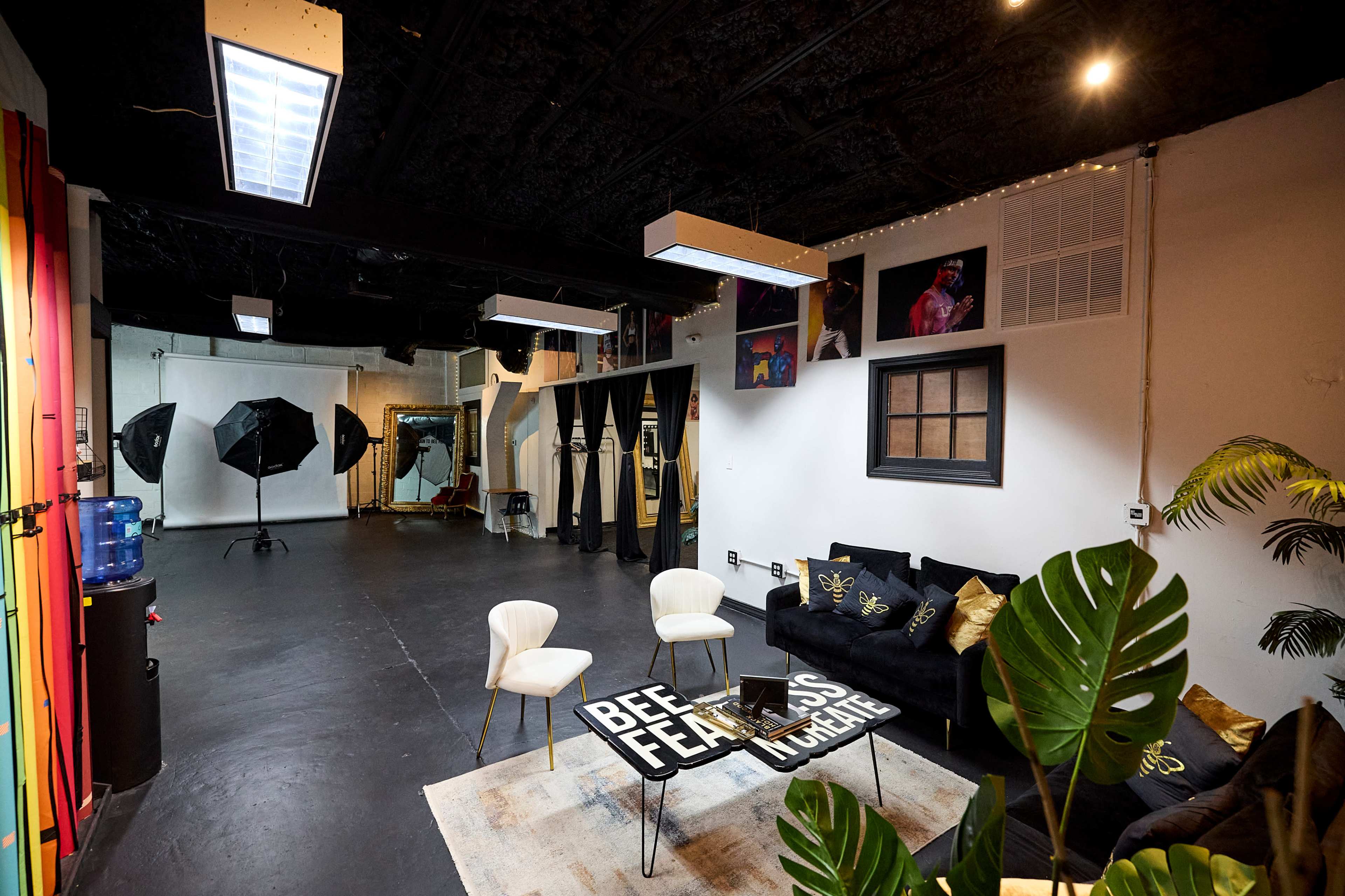 The image shows a spacious studio with black flooring, featuring a seating area with two chairs, a large rug, and various photography equipment in one corner.