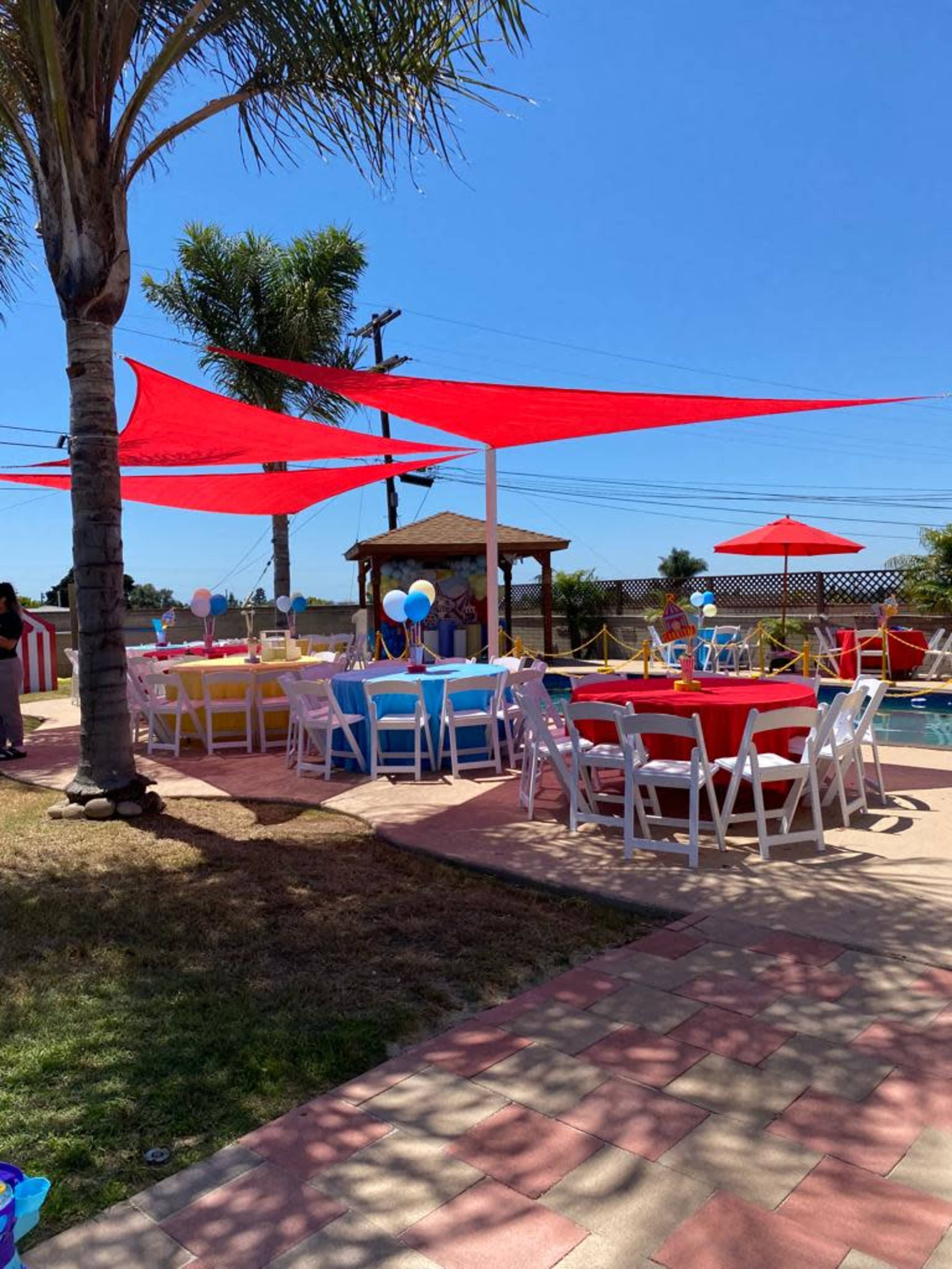 The image shows a vibrant outdoor space with colorful tables and chairs set up near a pool, surrounded by palm trees and decorative canopies.