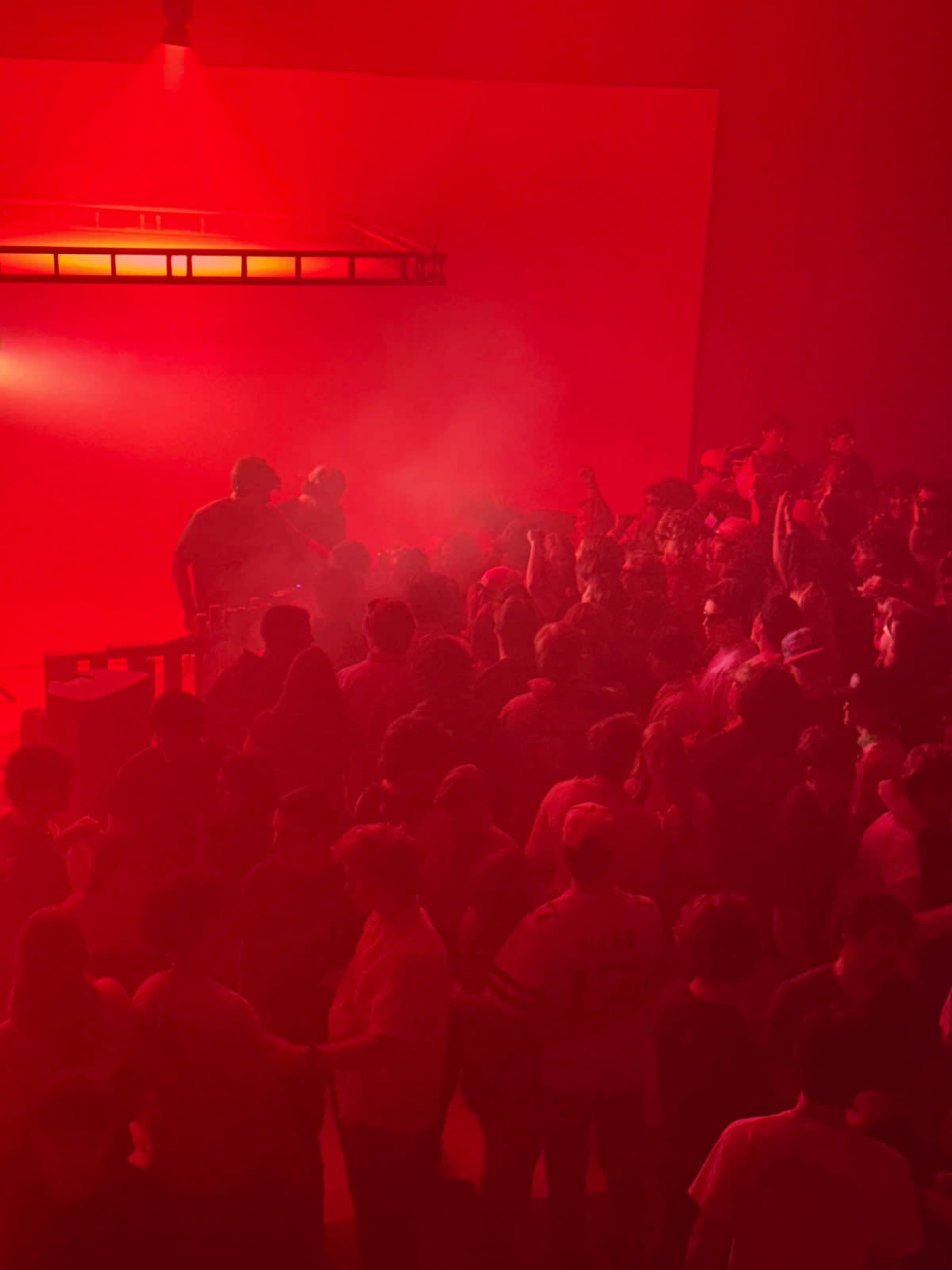 A dense crowd gathers under a red-lit atmosphere with fog as a DJ performs on stage.