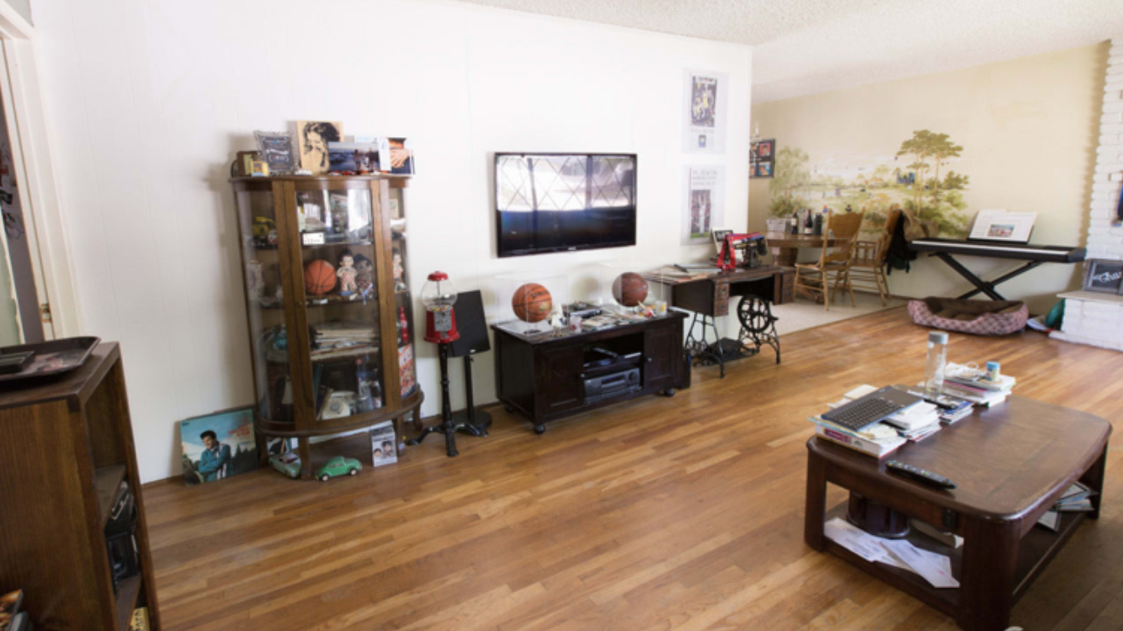 The living room features wooden flooring, a TV mounted on the wall, a glass display cabinet filled with sports memorabilia, and a dining area with a mural.
