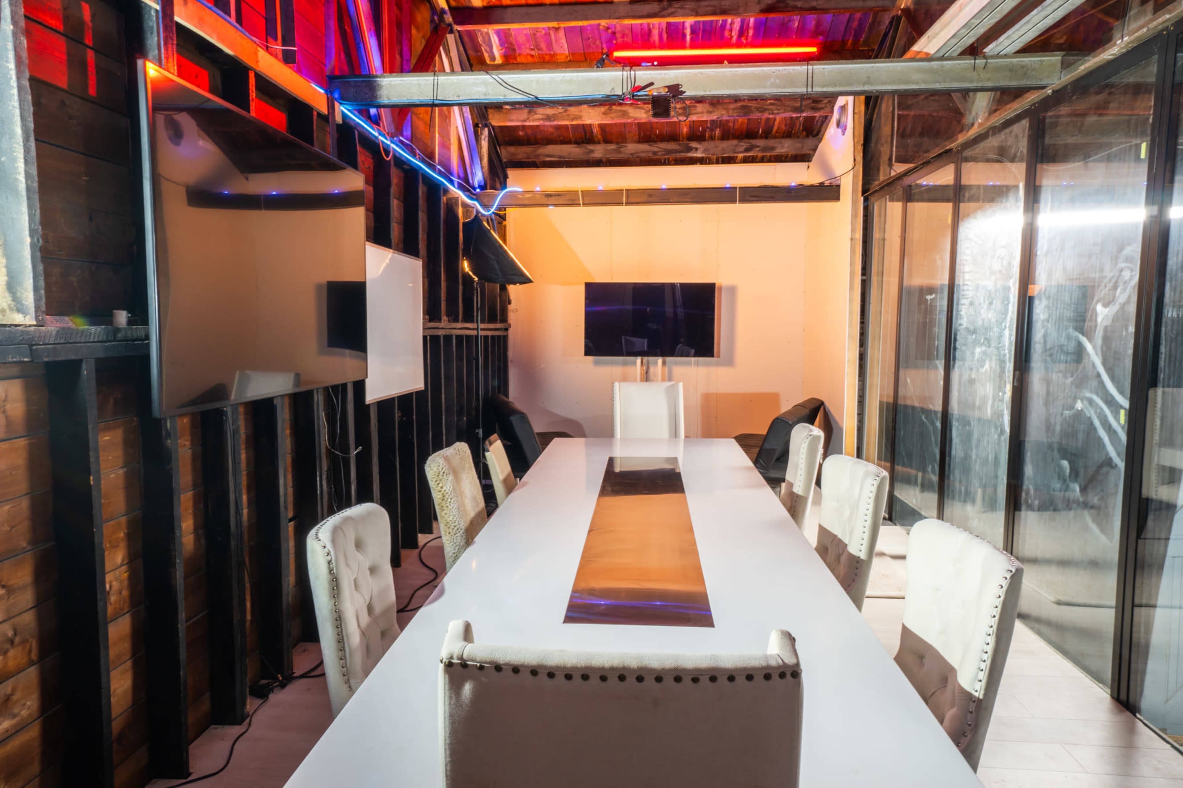 A long, white conference table is set in a dimly lit room with wooden walls and modern technology displayed on multiple screens.