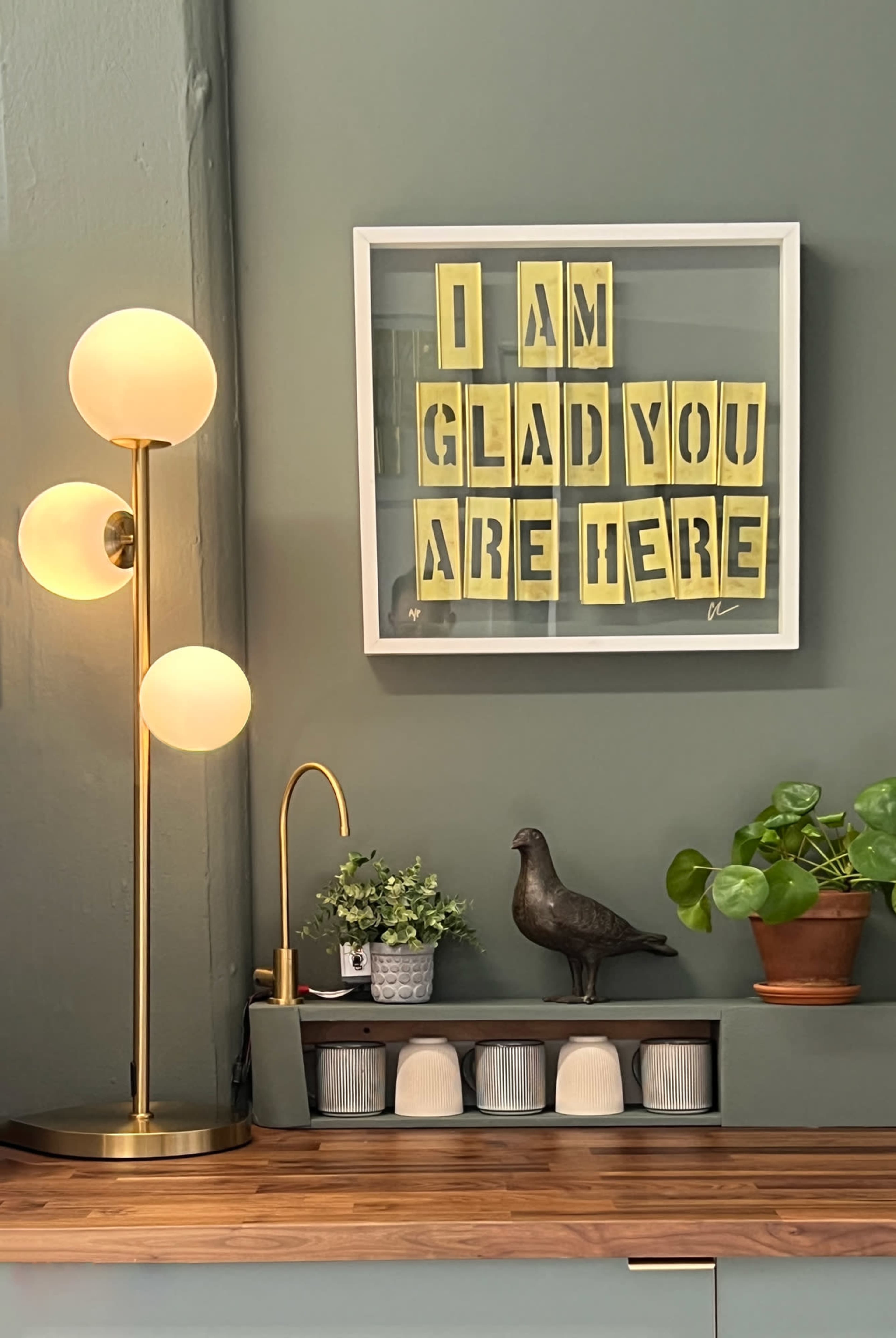 The image shows a framed artwork with yellow letters reading "I AM GLAD YOU ARE HERE" on a green wall, accompanied by a modern lamp, a decorative bird, and potted plants on a wooden countertop.