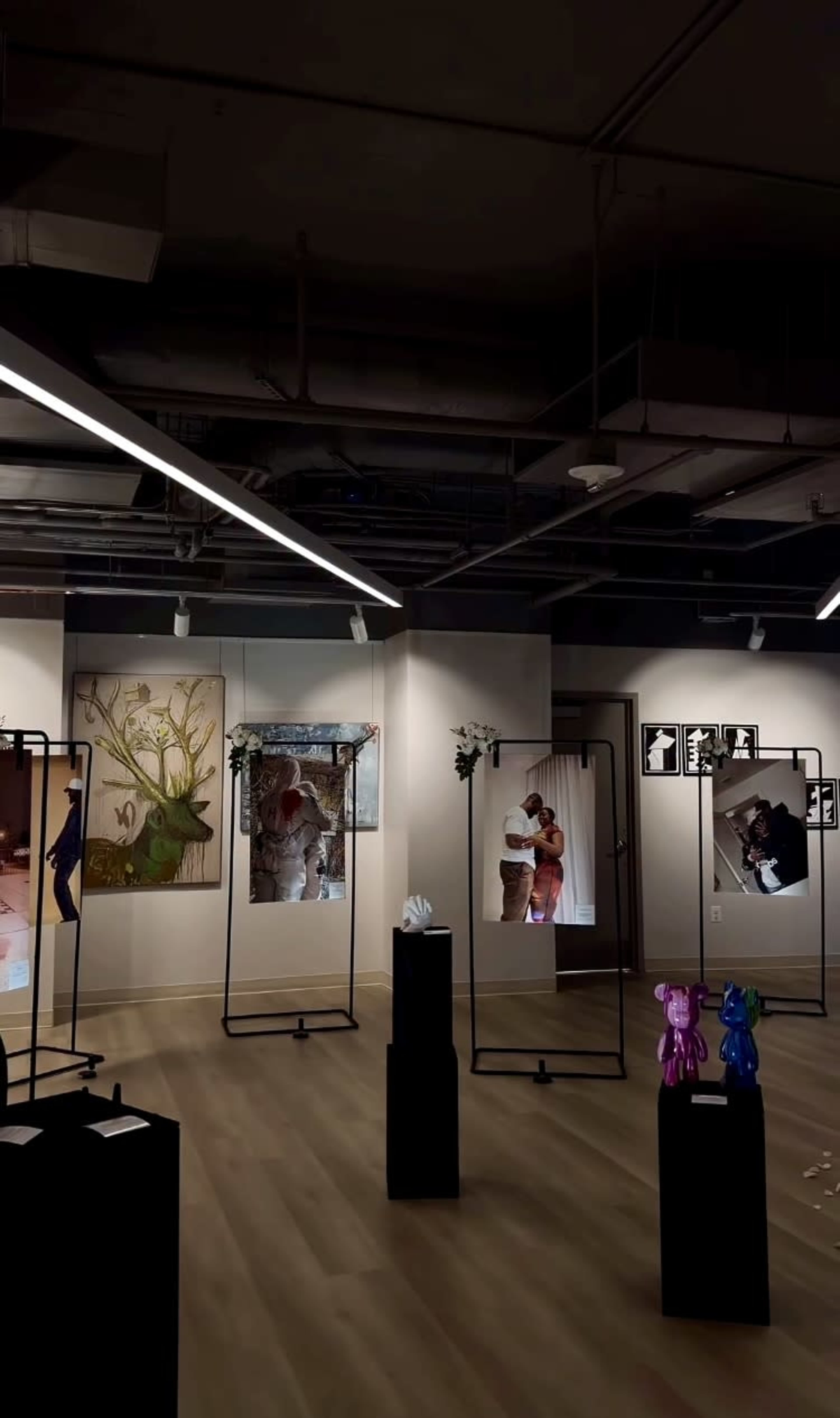 The image shows an art gallery featuring several framed artworks and sculptures displayed on pedestals, illuminated by overhead lighting.