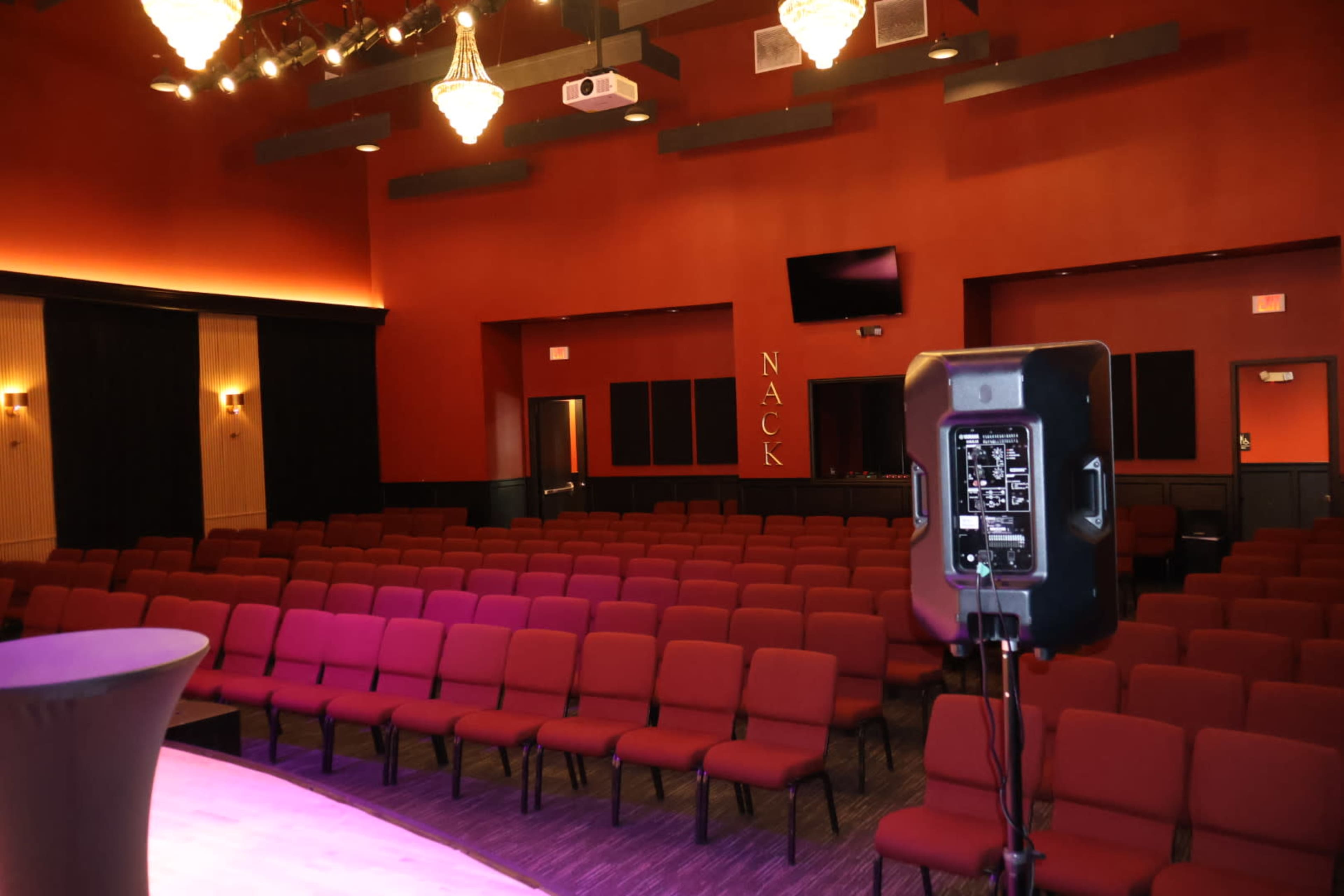 The image shows an interior view of a theatre with red walls, rows of red seats, and a stage with dim lighting.