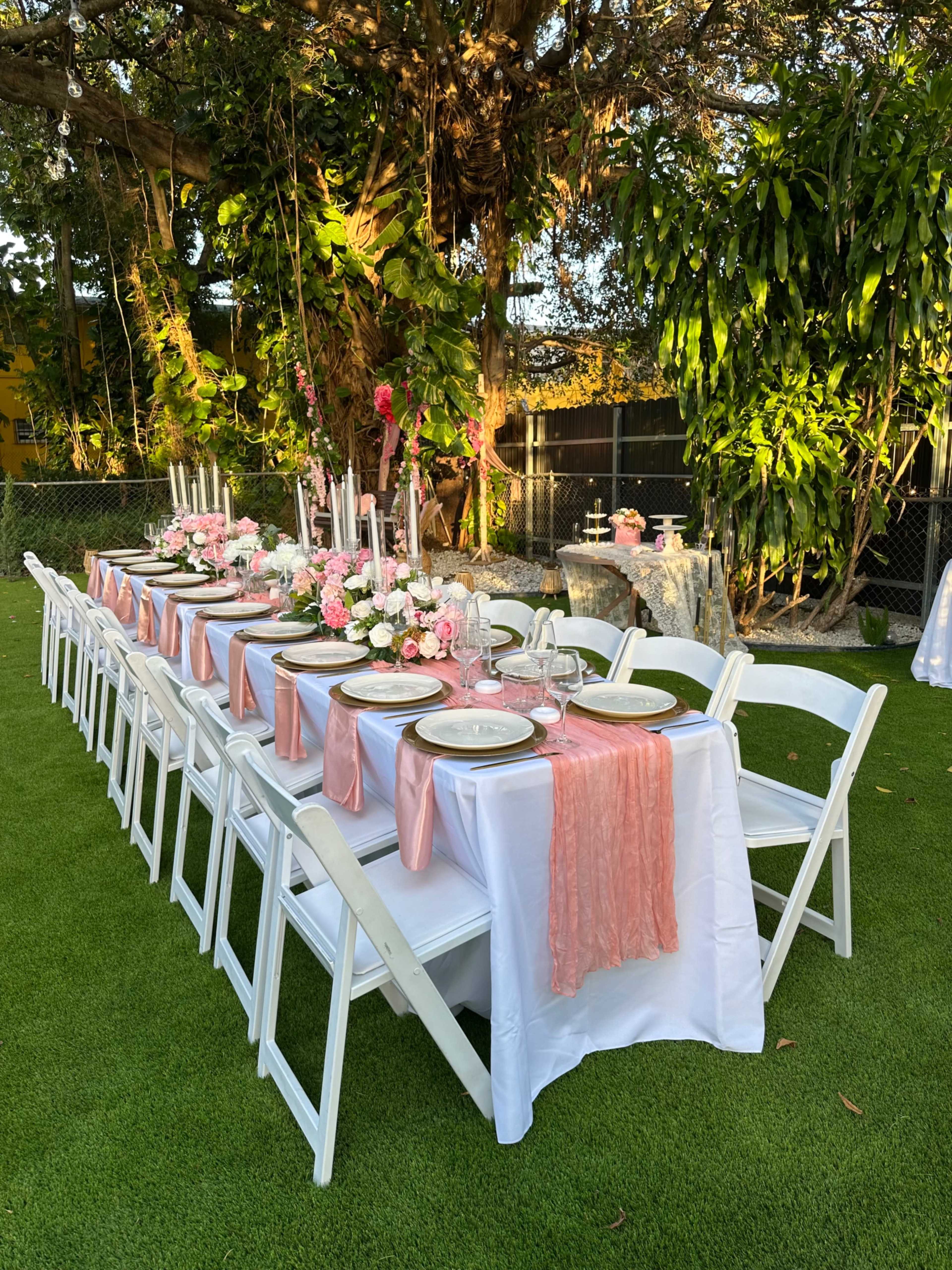 A long banquet table is elegantly set up outdoors, adorned with pink table runners, floral centerpieces, and candles, surrounded by lush greenery.