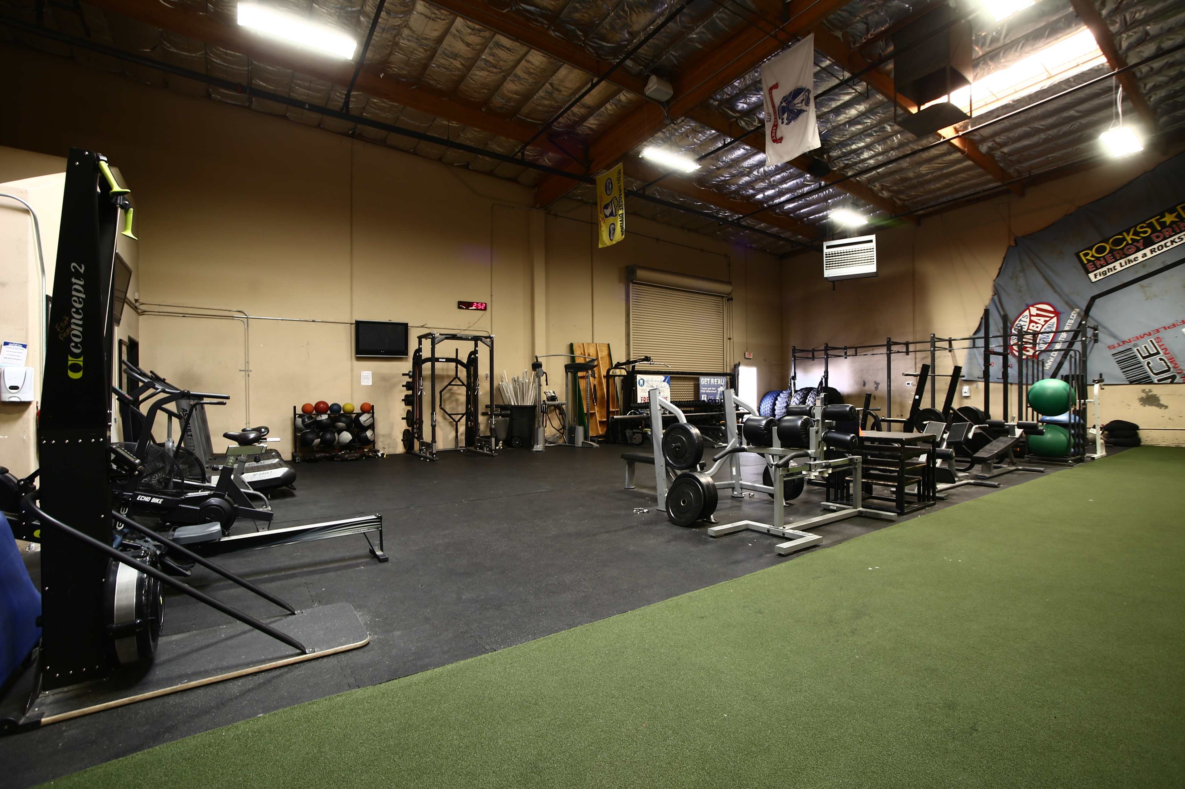 A spacious gym interior with various exercise equipment, including weight racks and cardio machines, against a backdrop of exposed metal beams and a turf area.
