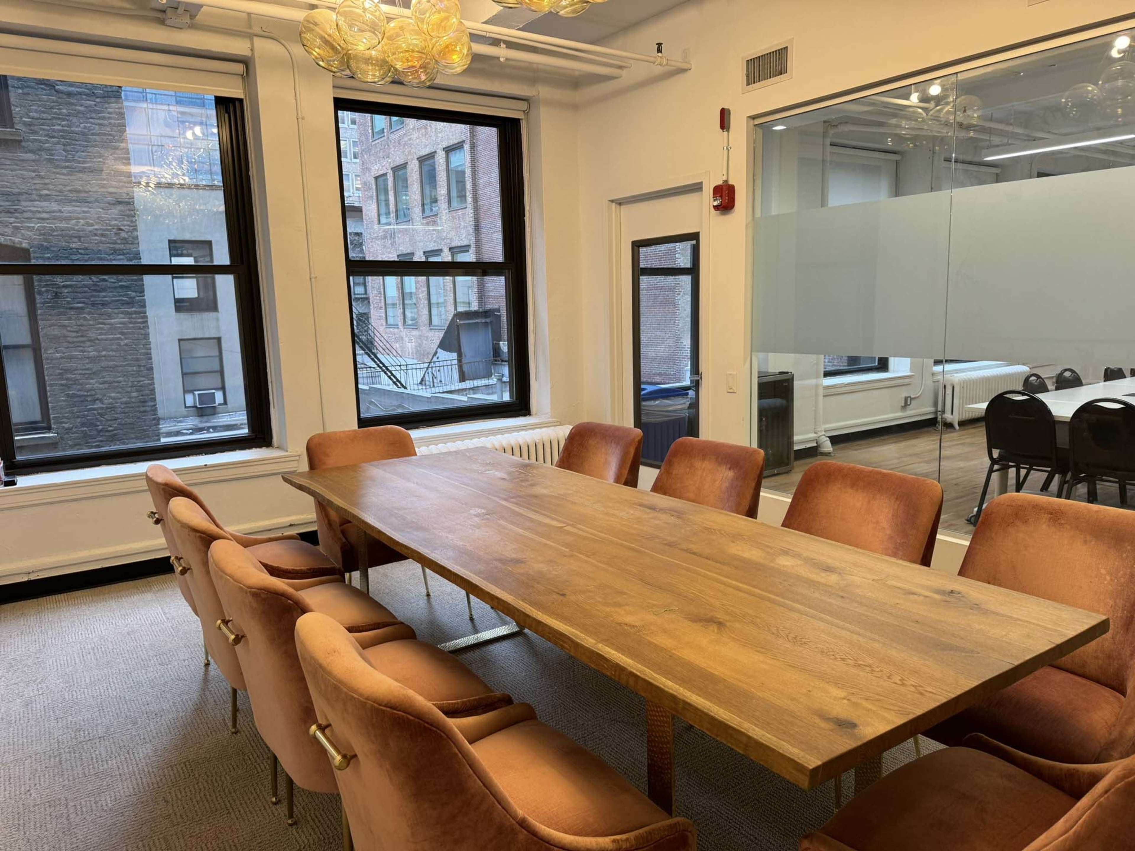 The image shows a conference room with a wooden table surrounded by upholstered chairs, large windows, and a glass wall.