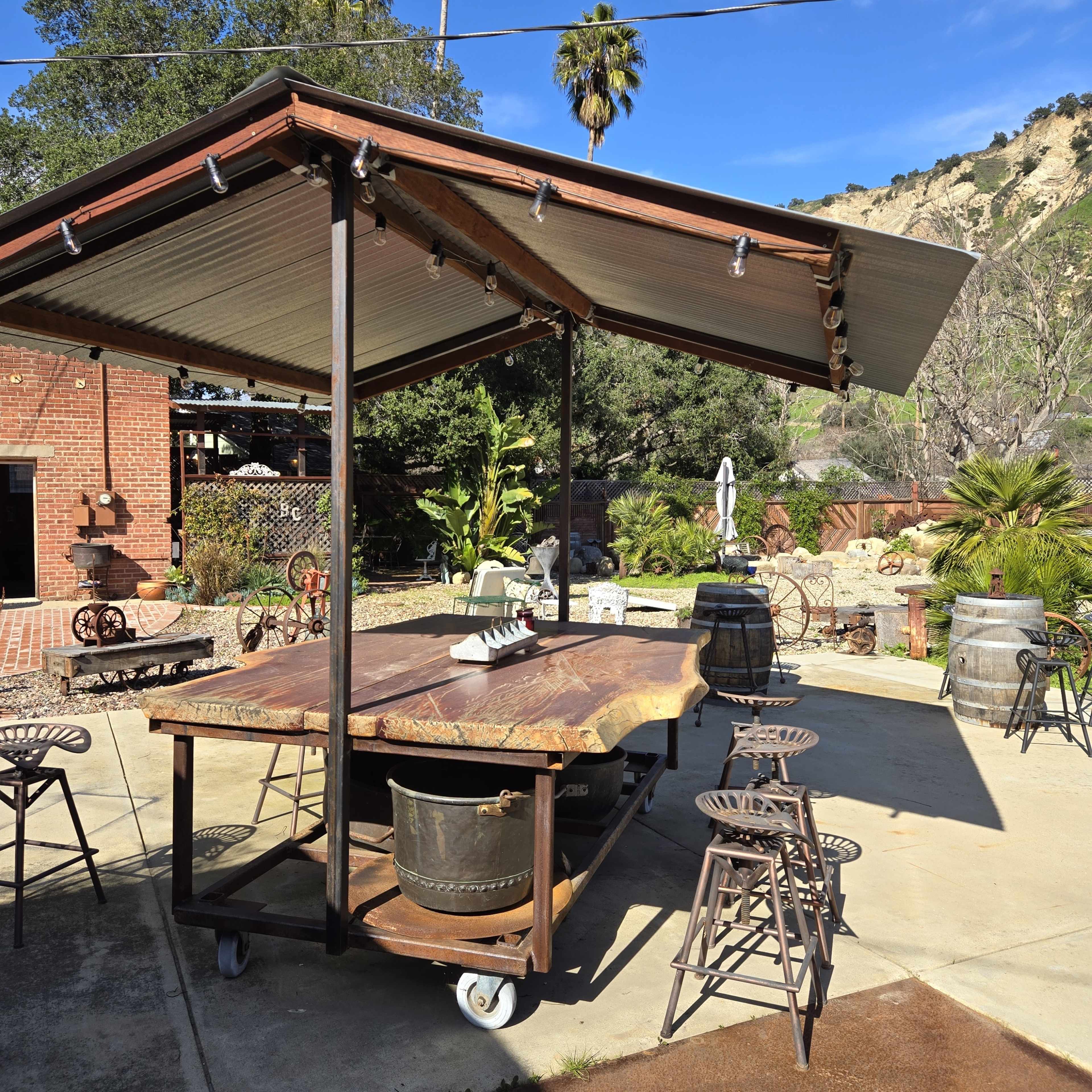 An outdoor seating area features a rustic wooden bar with metal stools under a slanted roof, surrounded by palm trees and landscaped greenery.