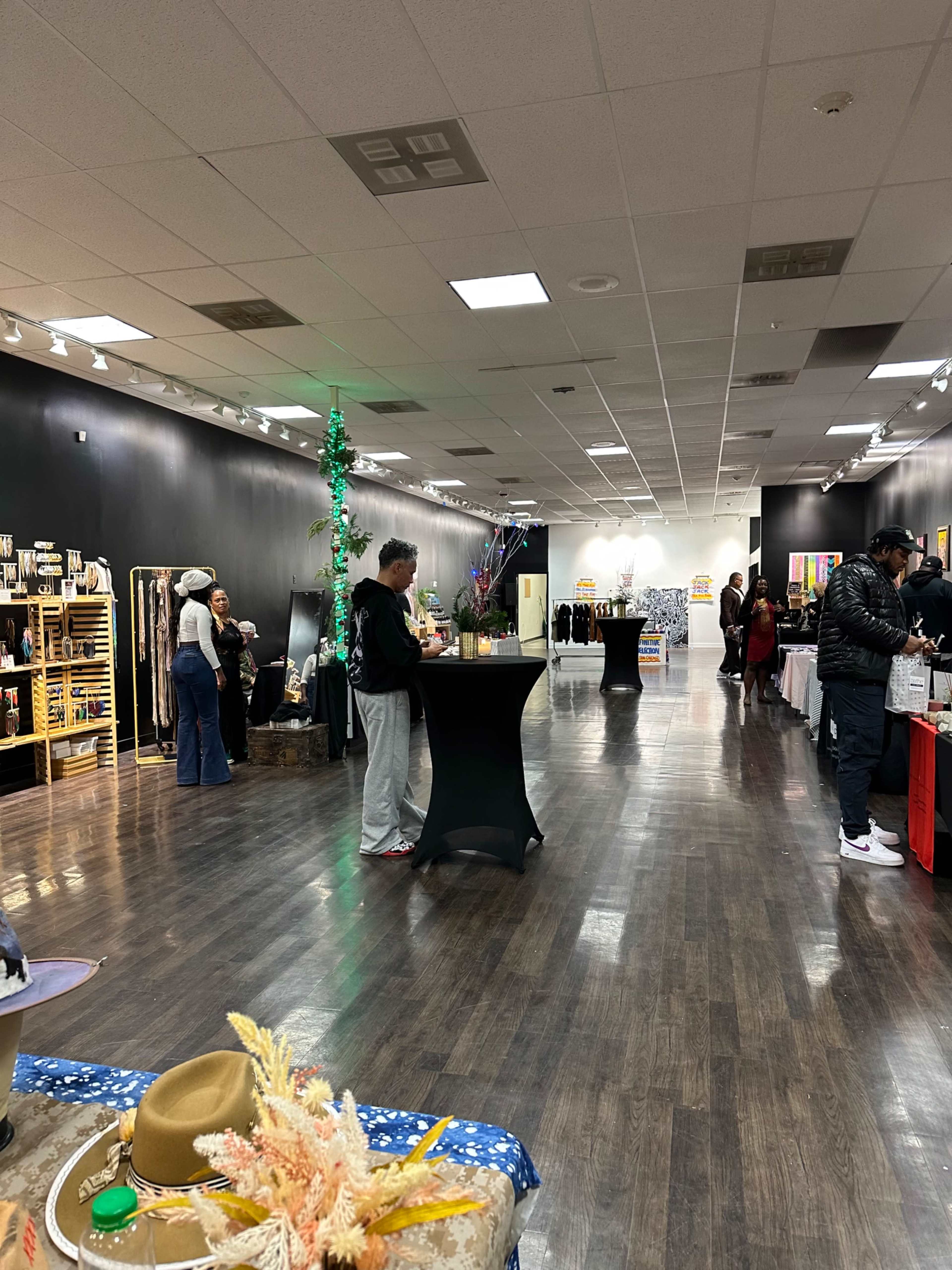 A spacious retail shop with black walls, featuring various vendors set up at tables along one side and a few individuals socializing in the center.
