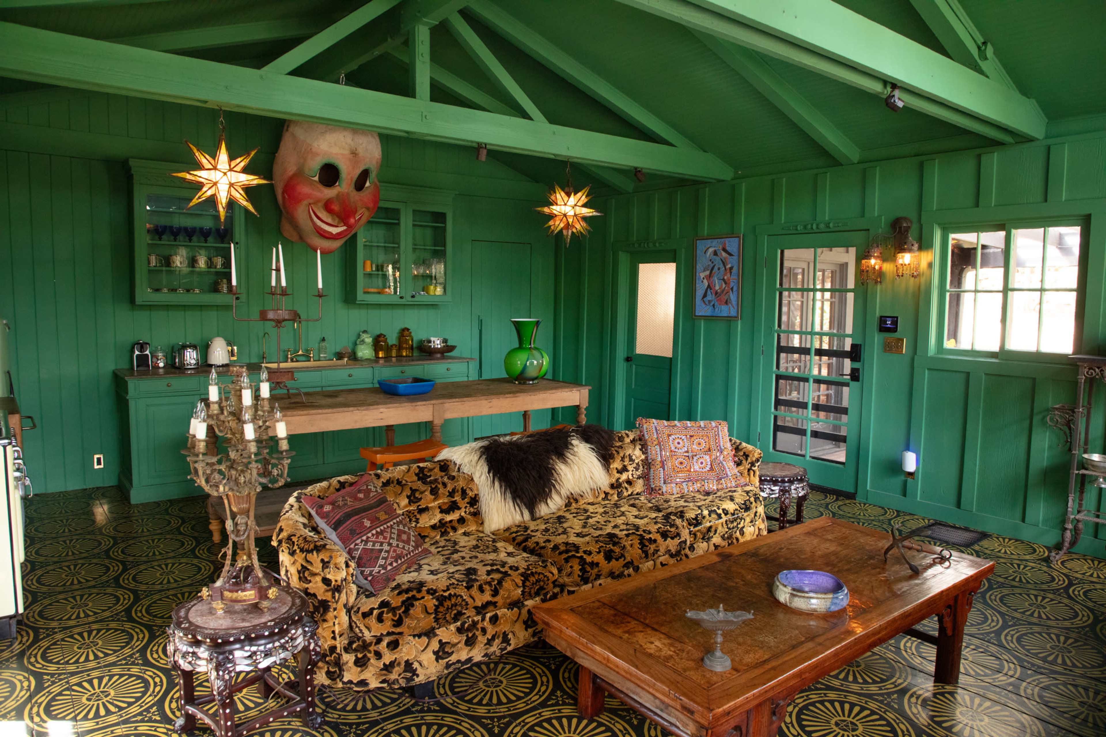 The image shows a vibrant green room with a plush black and gold sofa, a wooden coffee table, and a kitchen area featuring a large clown mask hanging on the wall.