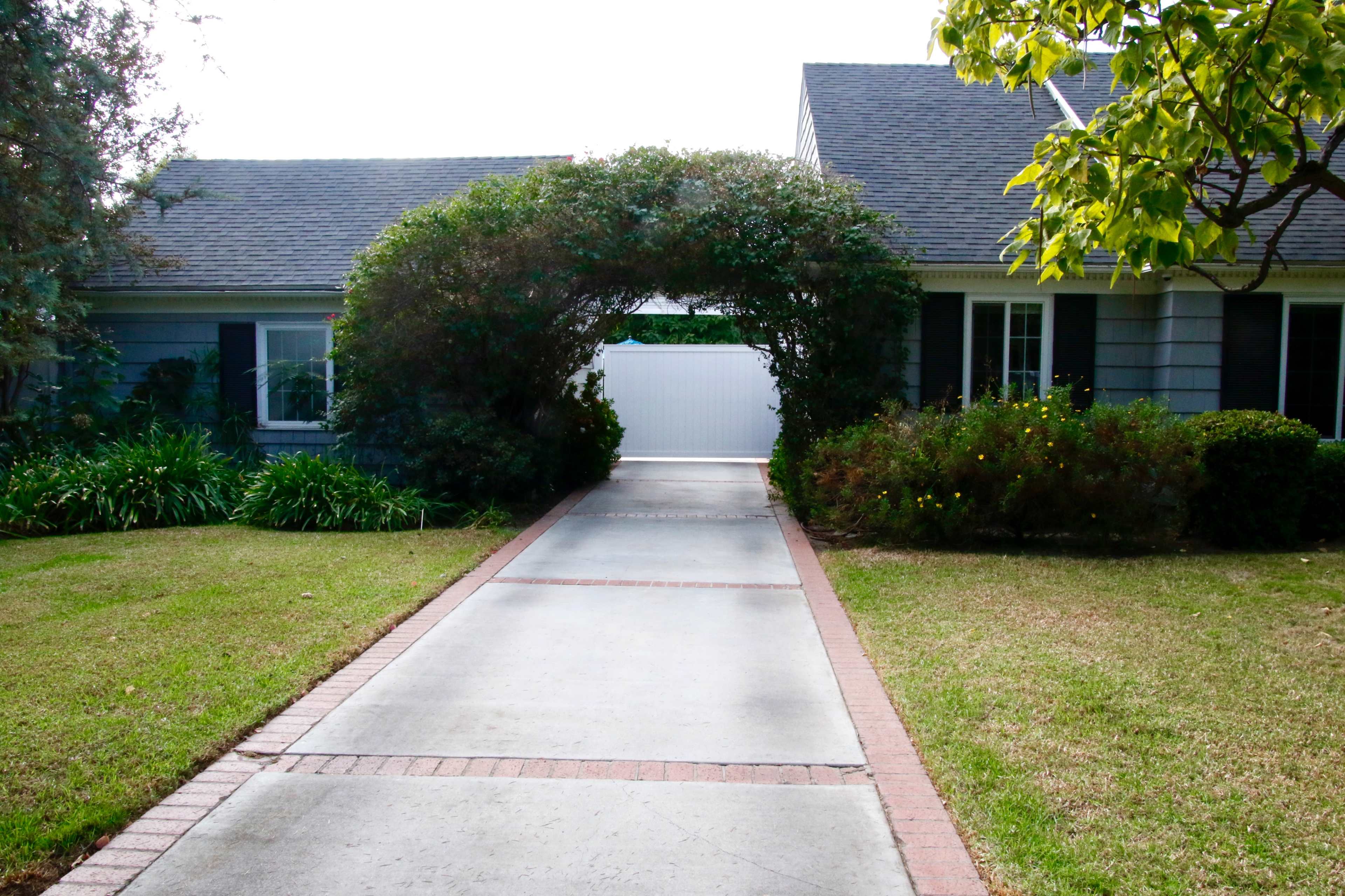 A pathway leads through an arched hedge toward a white gate between two houses.