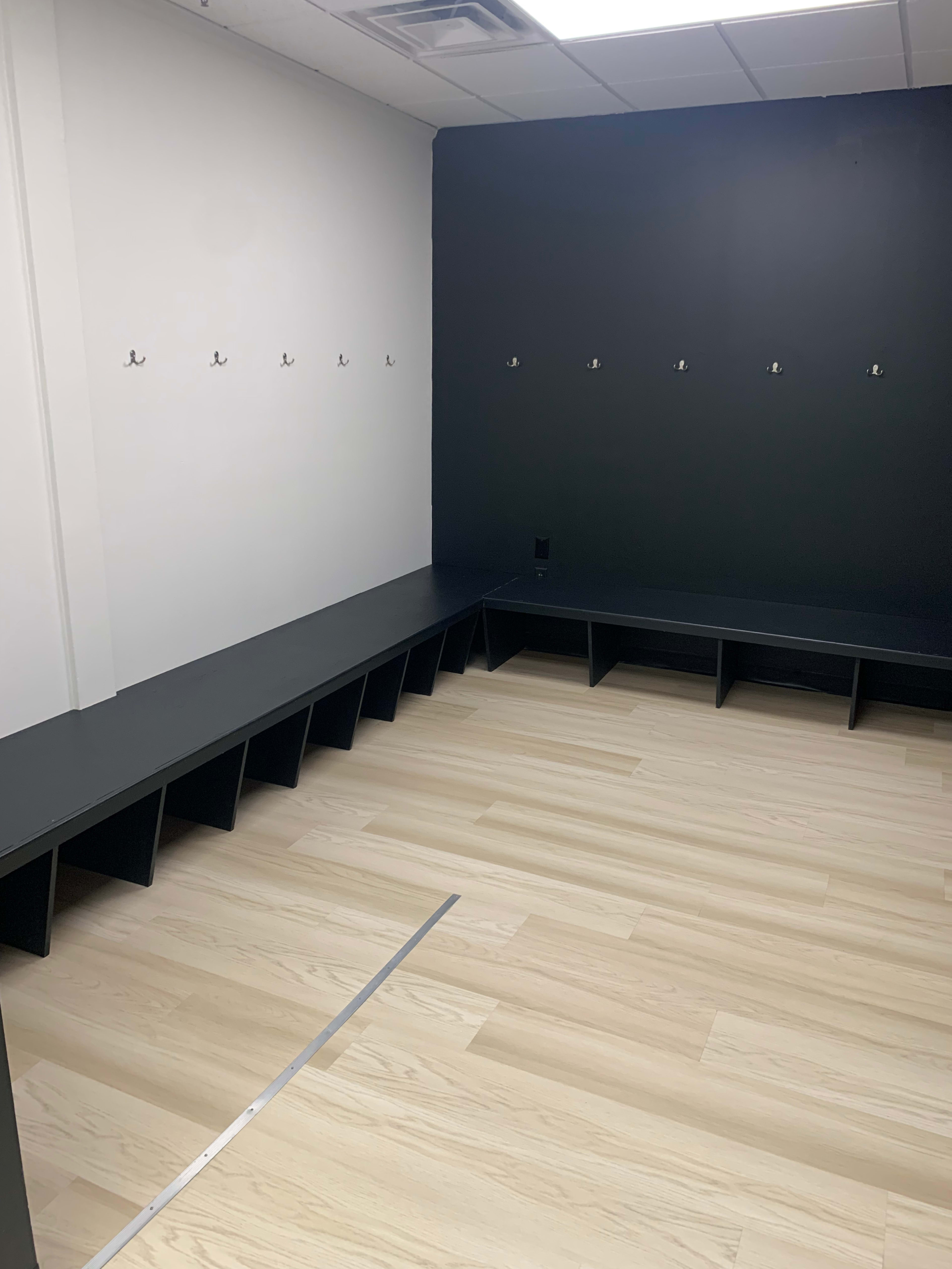 A sparsely furnished locker room with a dark wall, wooden floor, and several coat hooks mounted on the wall.