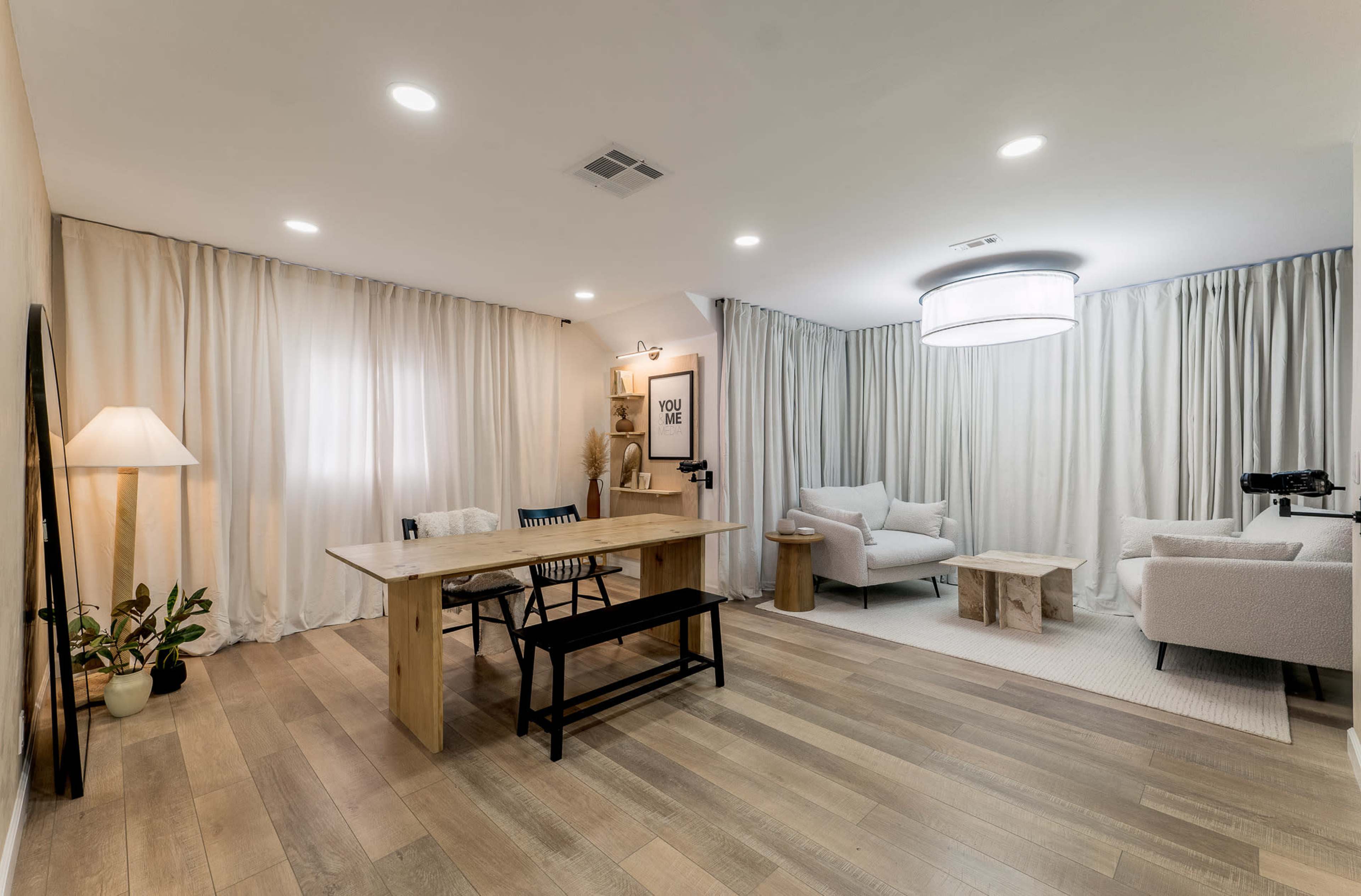 A modern living room featuring a wooden dining table and chairs, light-colored curtains, and minimalist furniture arranged around a central area.