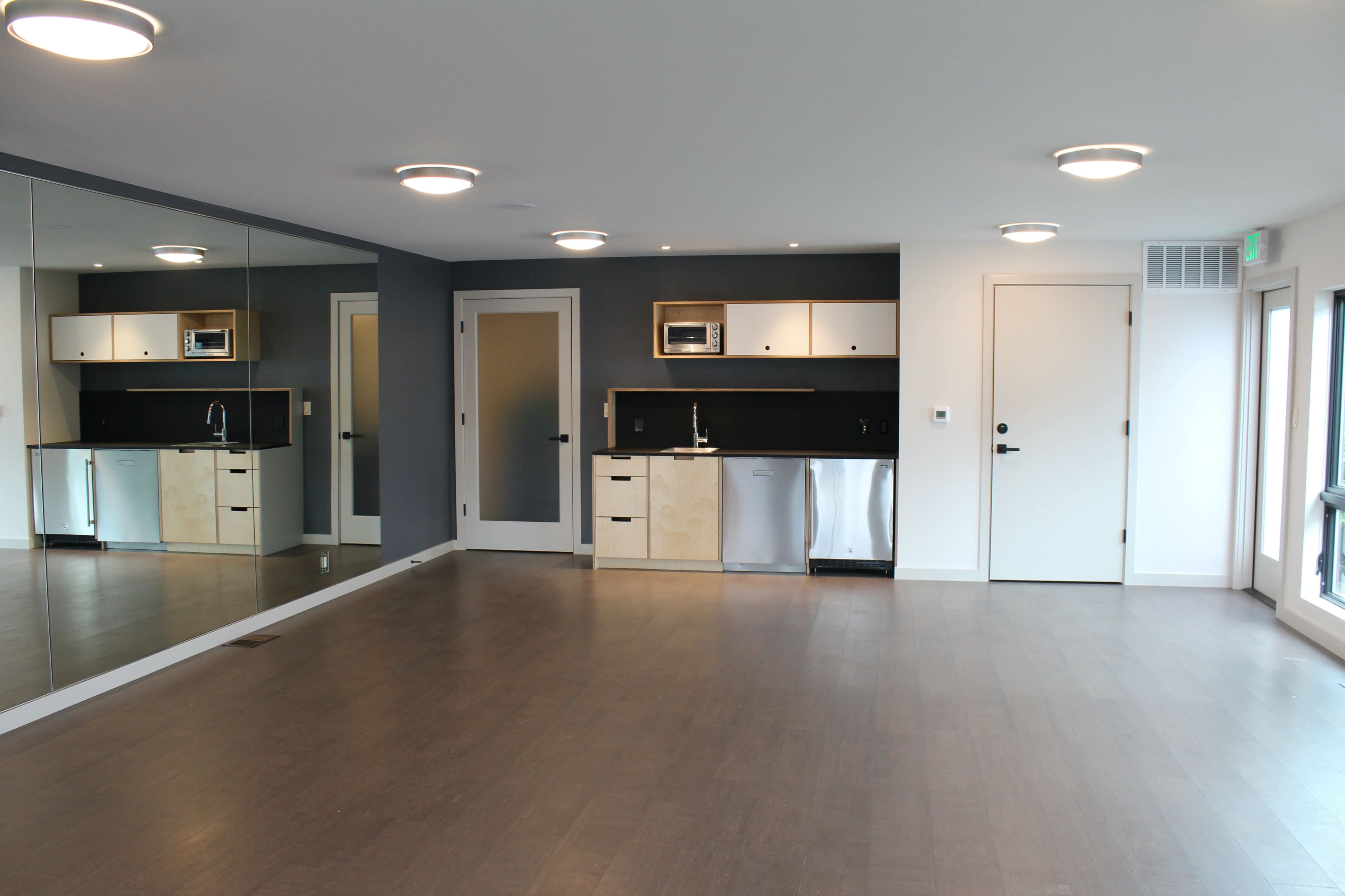 The image shows a modern, open living space with a kitchenette, large mirror on one wall, and minimalist design elements.
